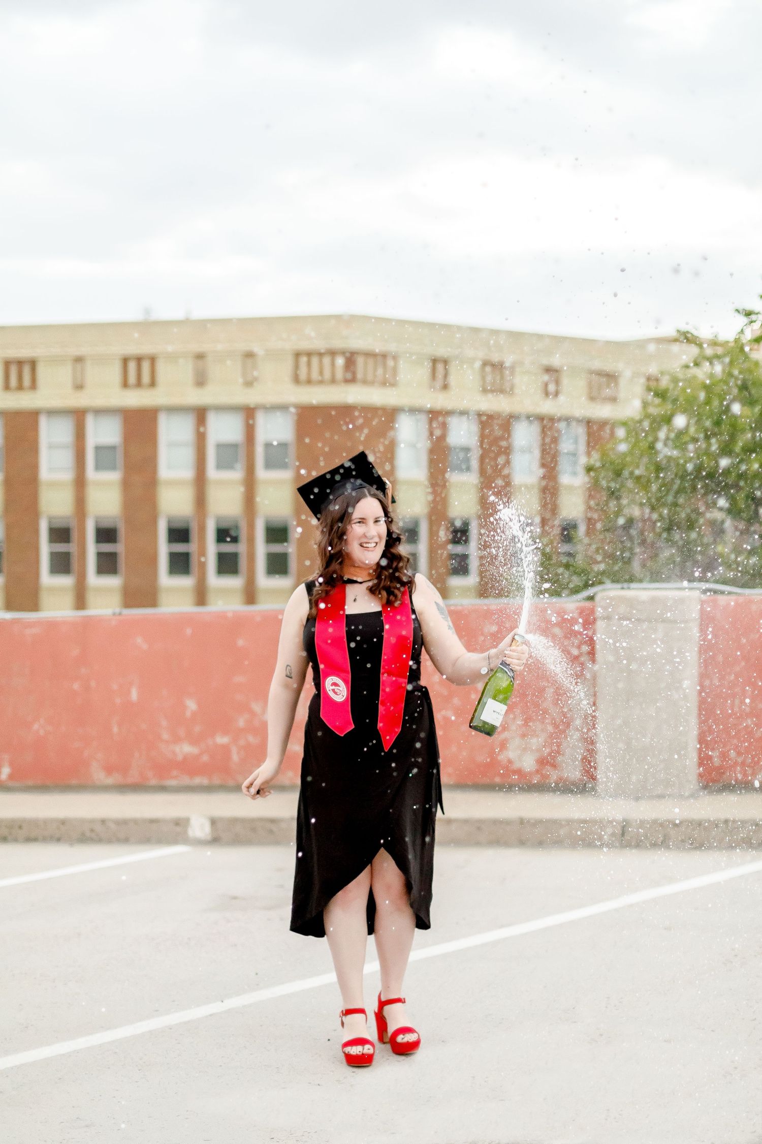 Smiling graduate in black dress and cap popping champagne with confetti on a rooftop parking lot.