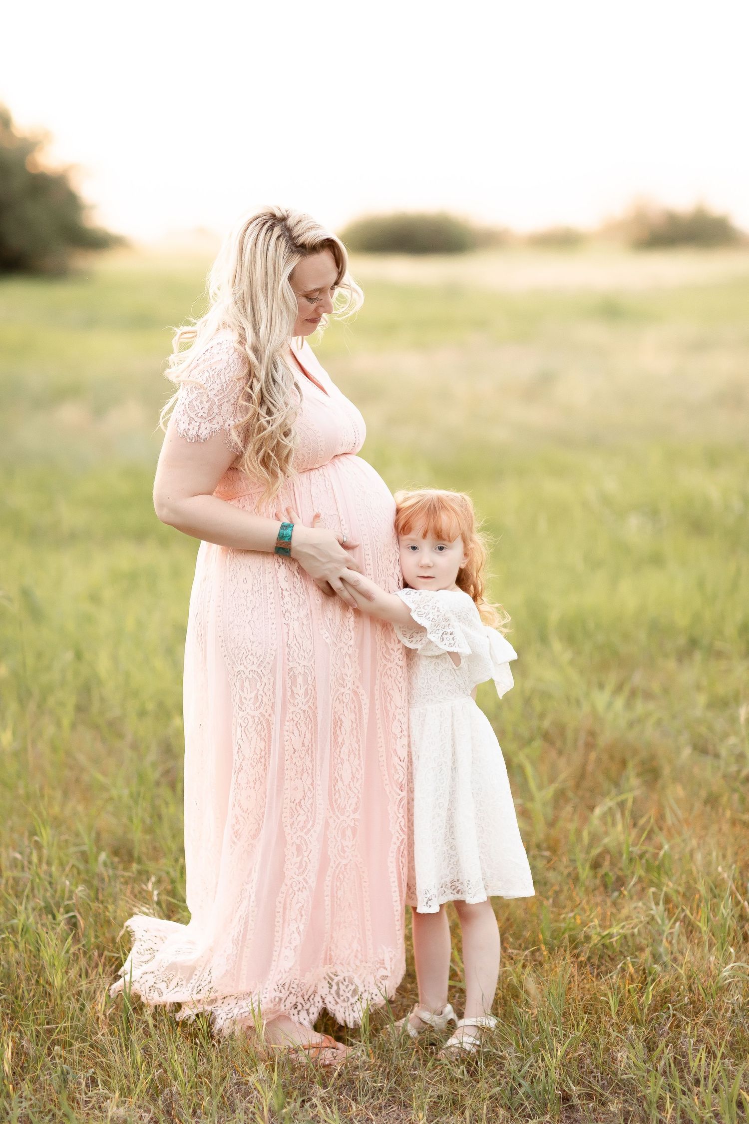 Pregnant woman in pink lace gown with redhead daughter hugging her baby bump in a sunlit field.