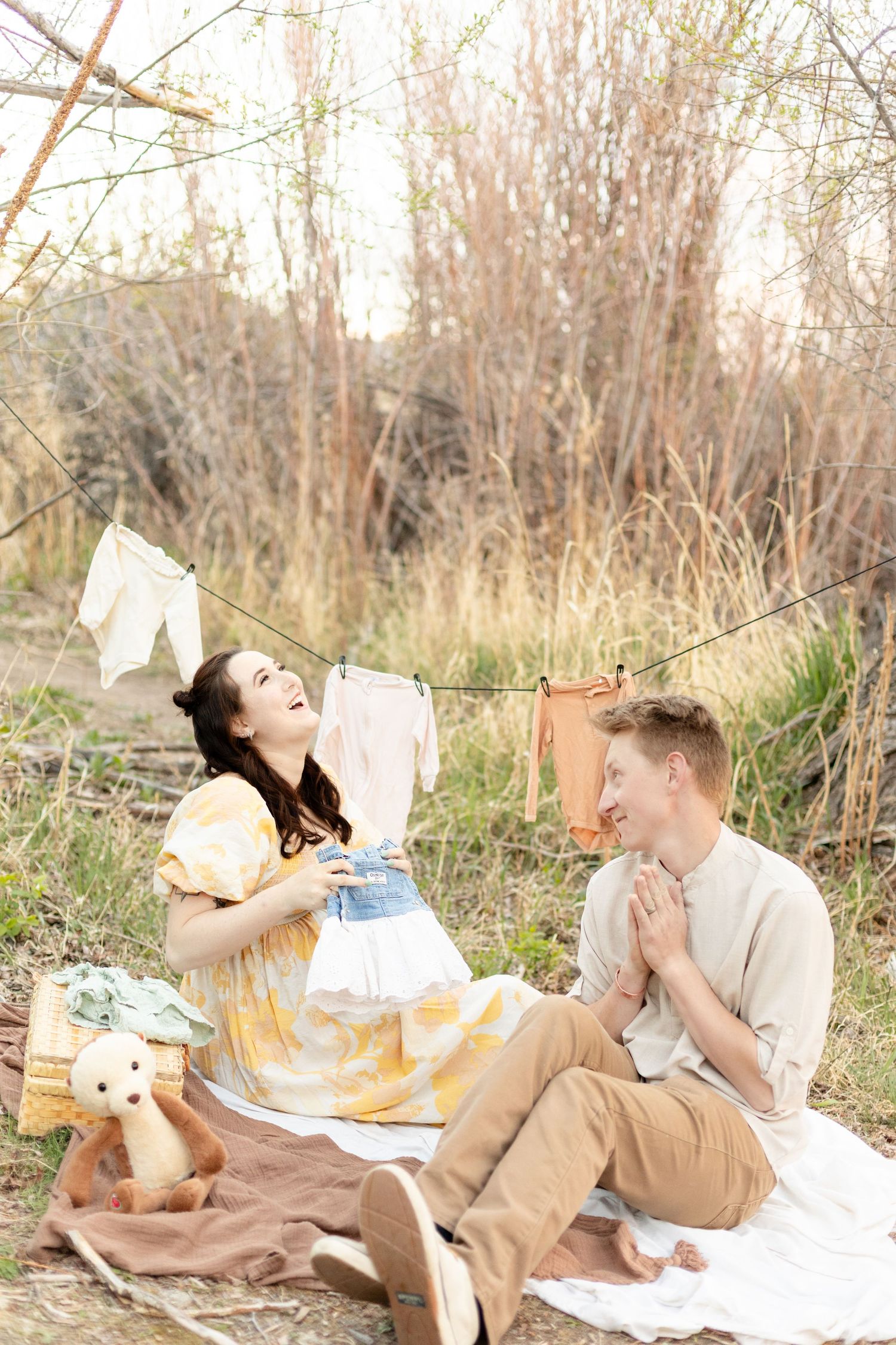 Laughing pregnant woman in yellow floral dress sits with partner on blanket near baby clothesline outdoors.