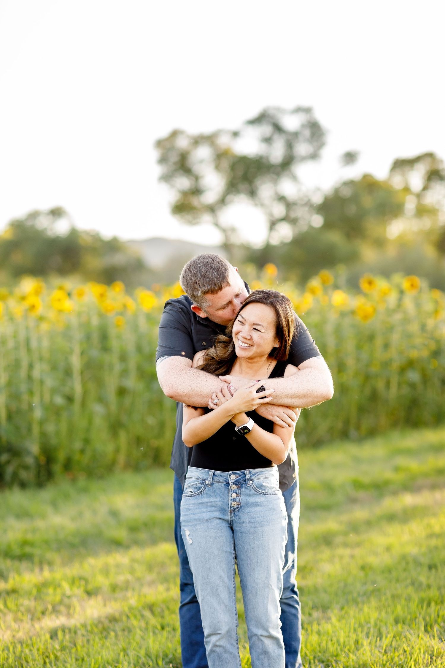 Man in black shirt hugging woman from behind in front of a sunflower field during golden hour engagement photos.