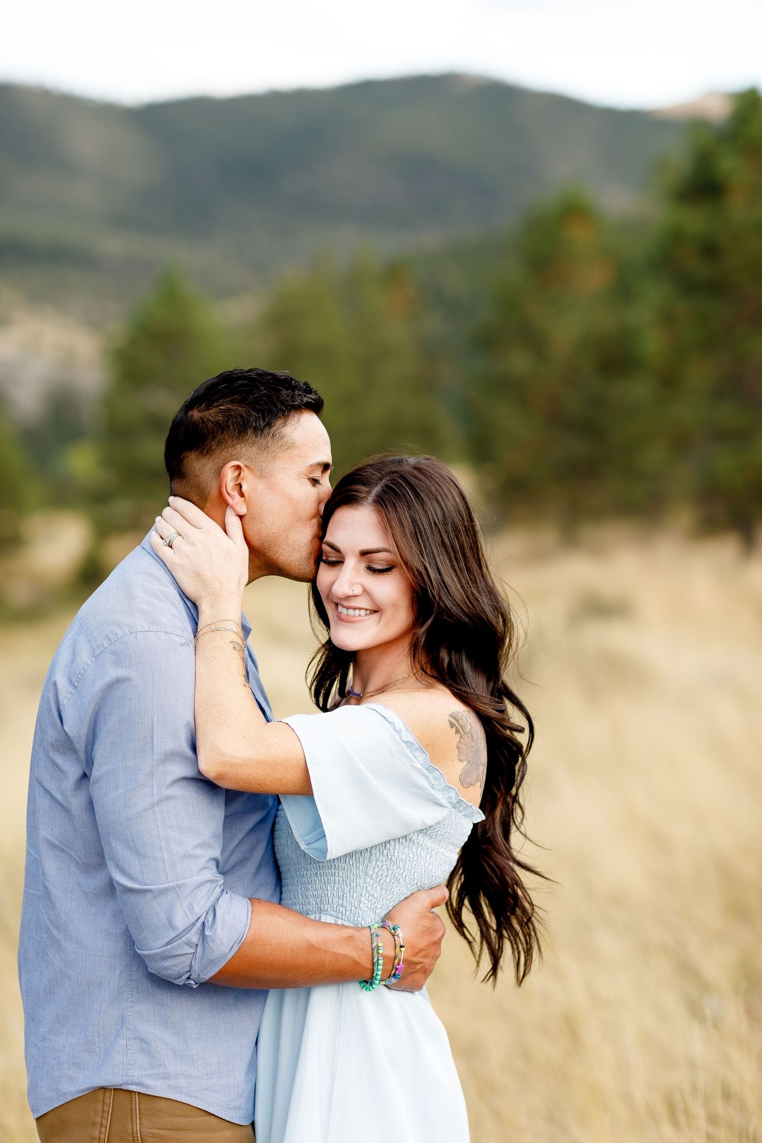 Man kissing woman on the cheek as she smiles in a light blue dress during a mountain engagement session.