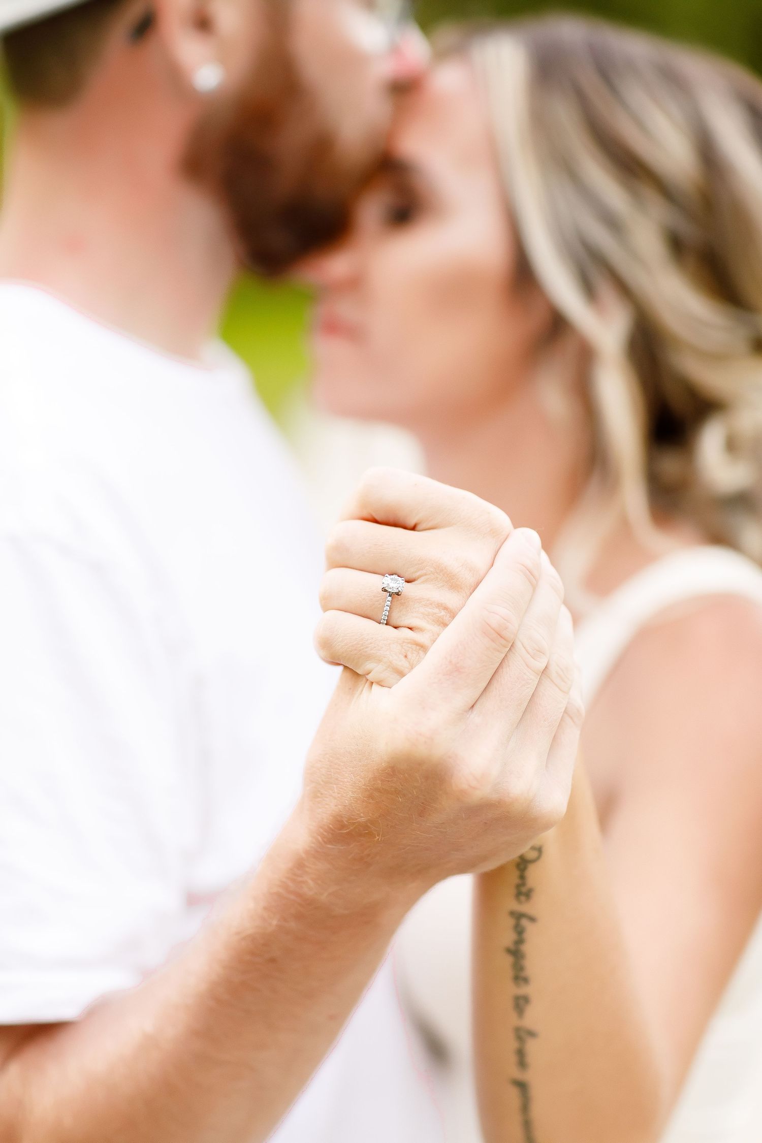 Close-up of an engagement ring on a woman's hand as couple shares a kiss in a soft bokeh background.