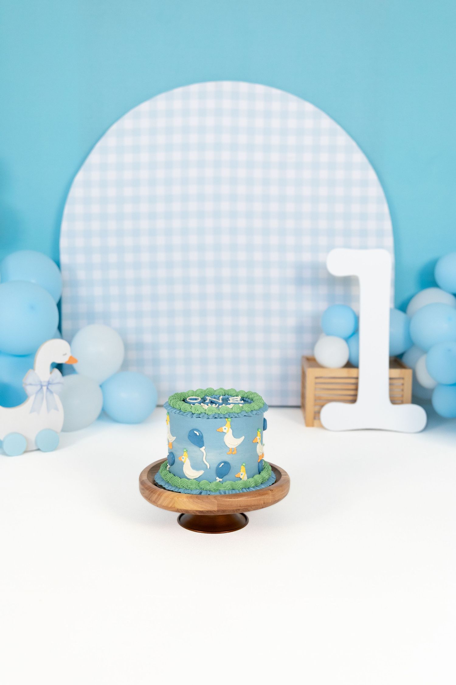 Blue duck-themed first birthday smash cake on wooden stand with gingham backdrop and balloon decorations.
