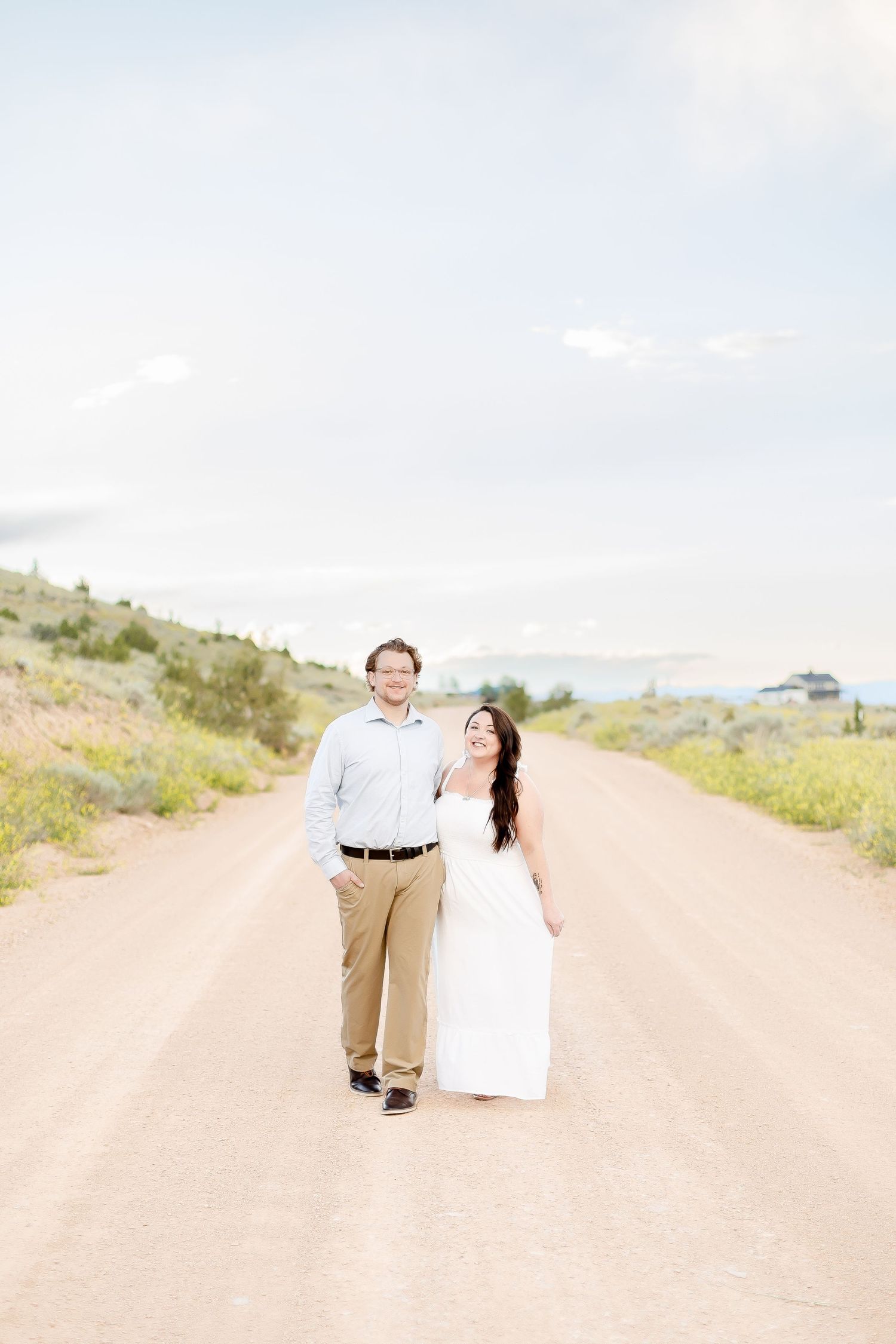 Smiling couple walking on a rural dirt road, woman in white dress looking up at man in light blue shirt.