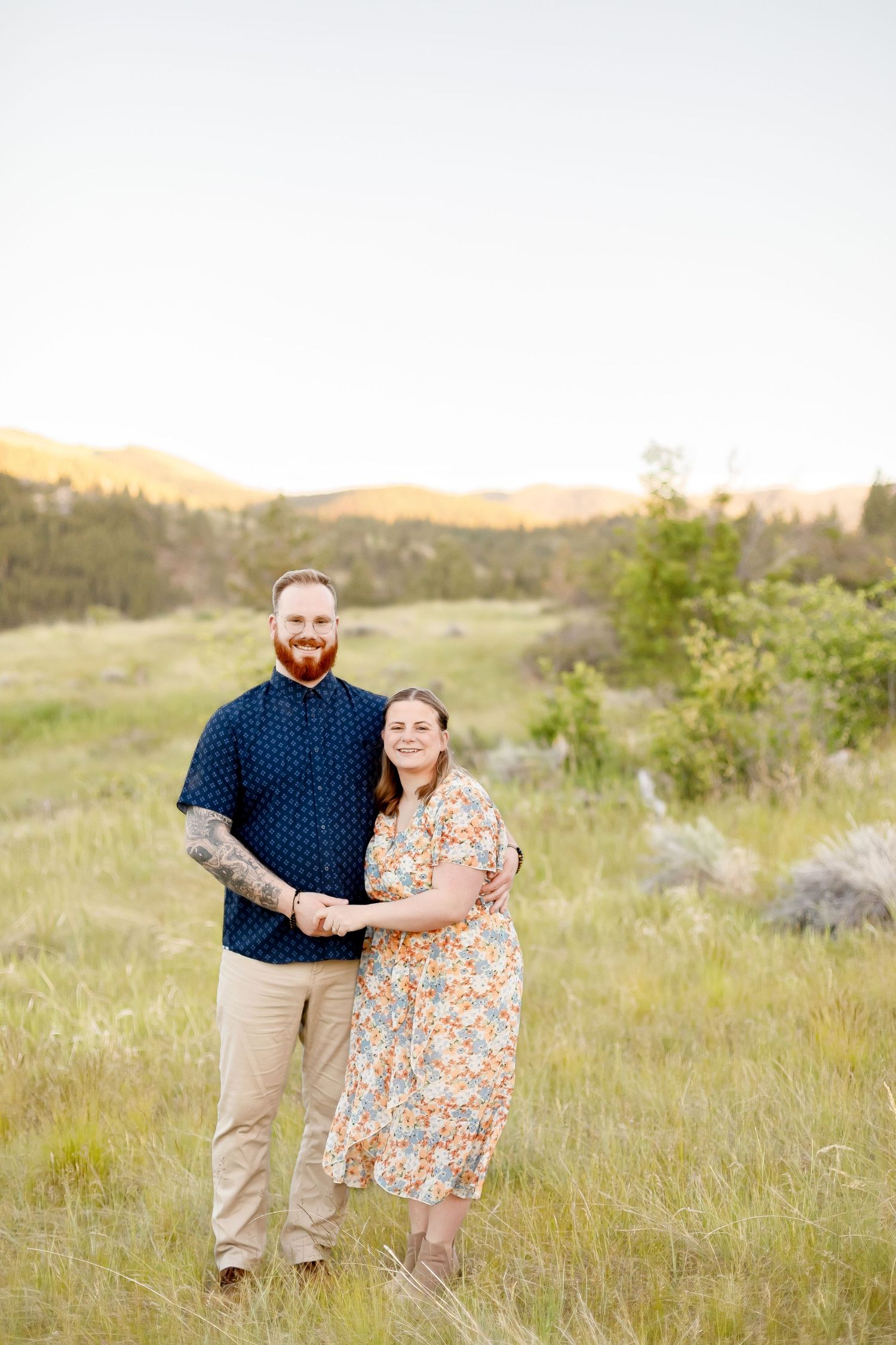 Couple standing in green meadow at sunset, man with red beard in navy shirt, woman in floral dress.
