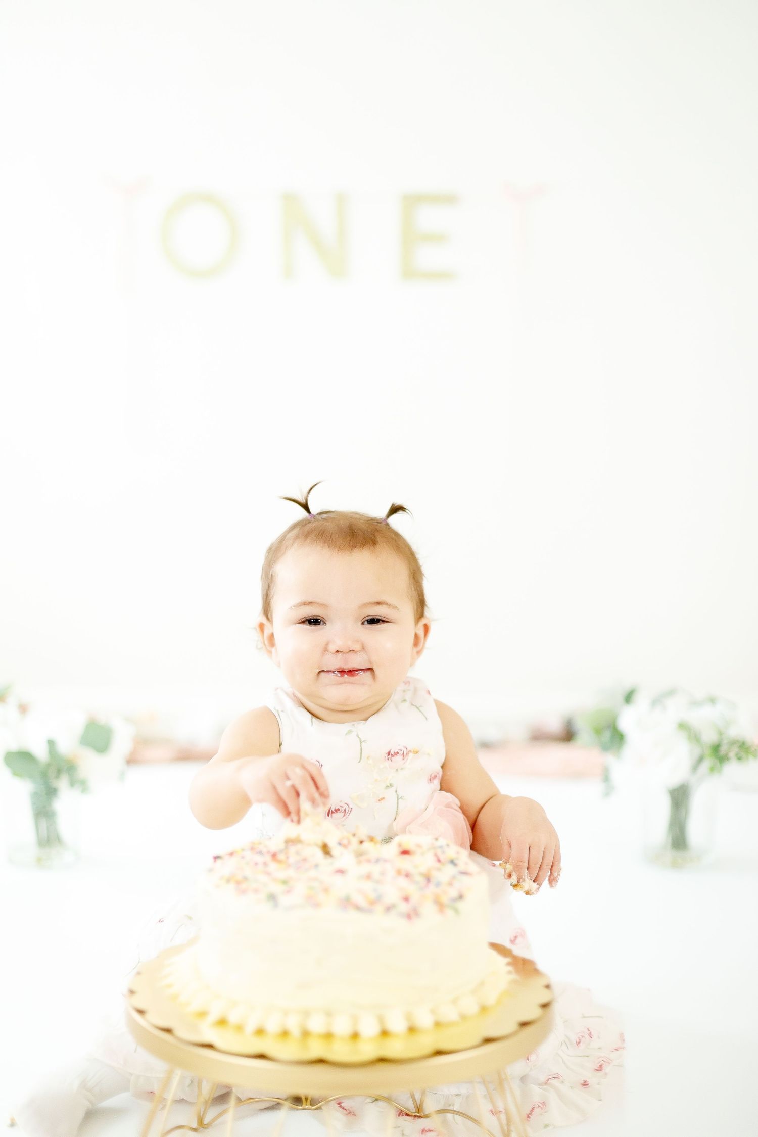 Smiling baby girl in pink floral dress touching white sprinkle cake at first birthday party.