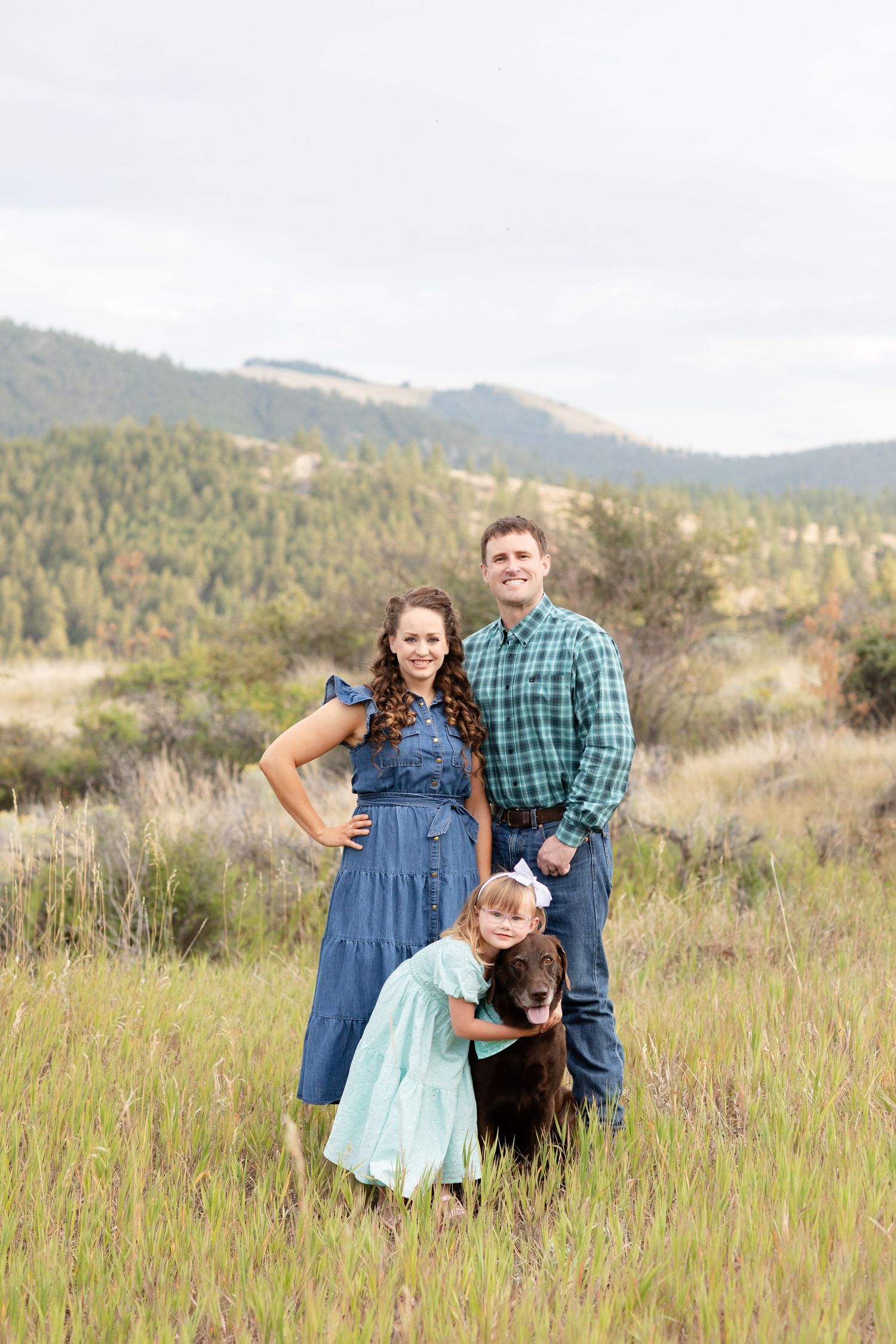 A young family with a chocolate Labrador poses in a lush green mountain meadow on a cloudy day.