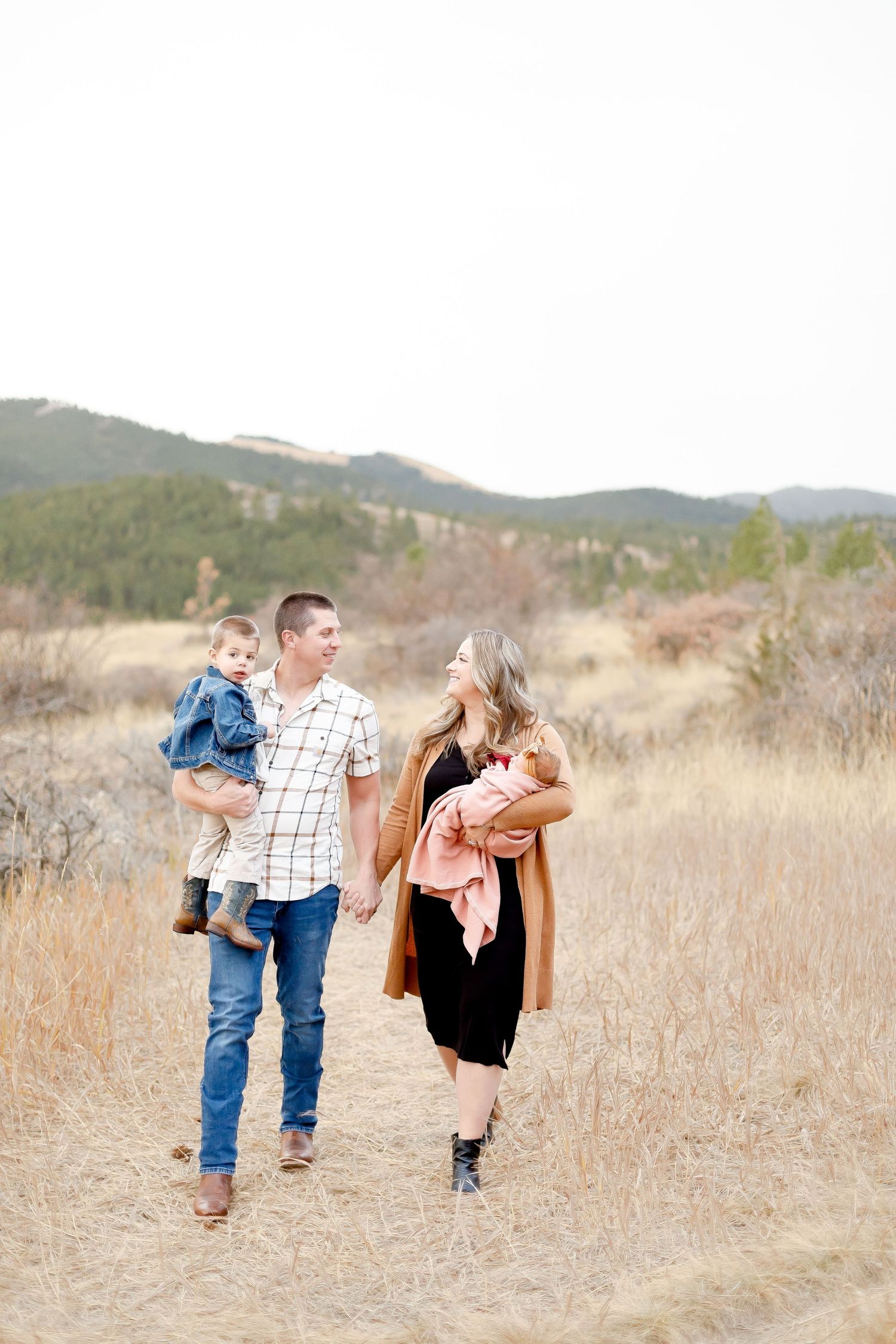 Young family of four walking hand in hand through dry grass meadow with mountain backdrop.