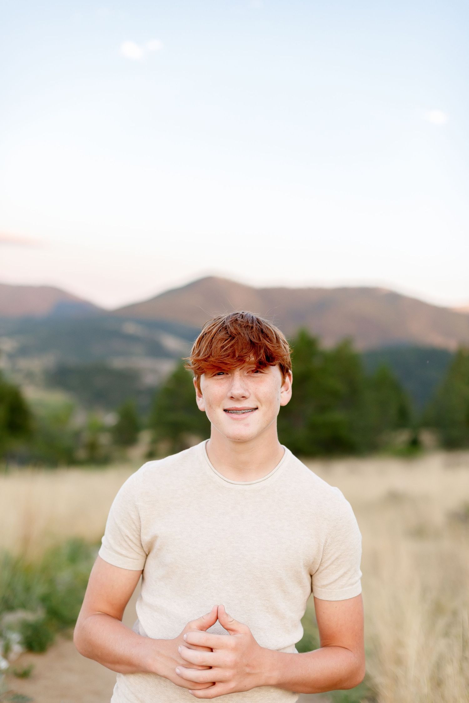 Smiling teenage boy with red hair wearing a beige t-shirt standing in a mountain meadow.