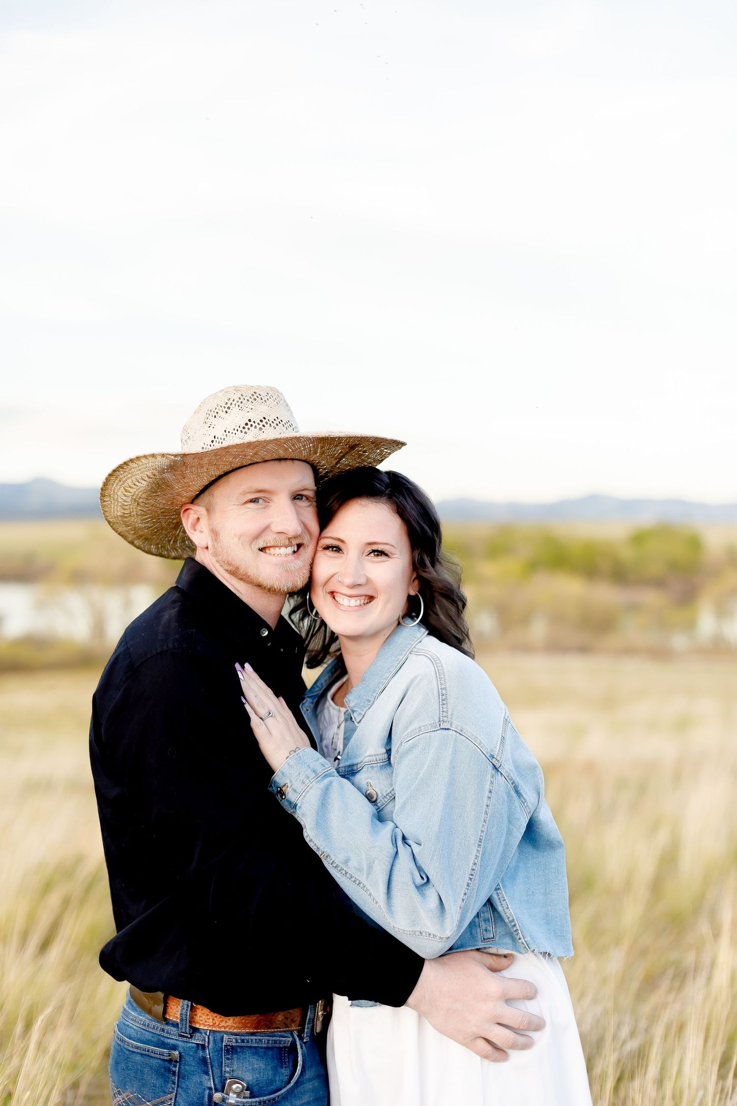 Smiling couple hugging in grassy field, man wearing straw cowboy hat and black shirt, woman in denim jacket.