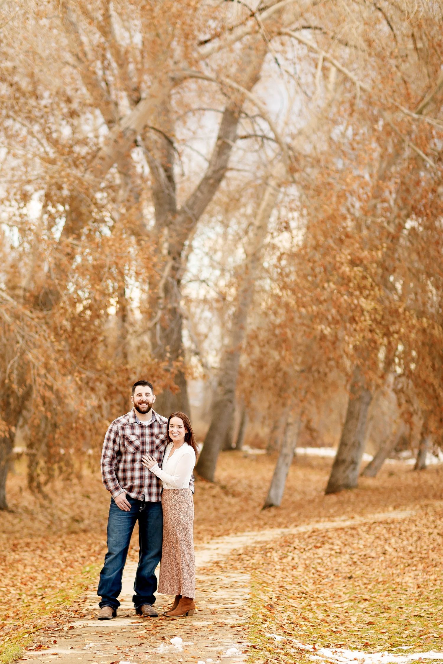 Couple posing on a leaf-covered path surrounded by golden autumn trees in a park.