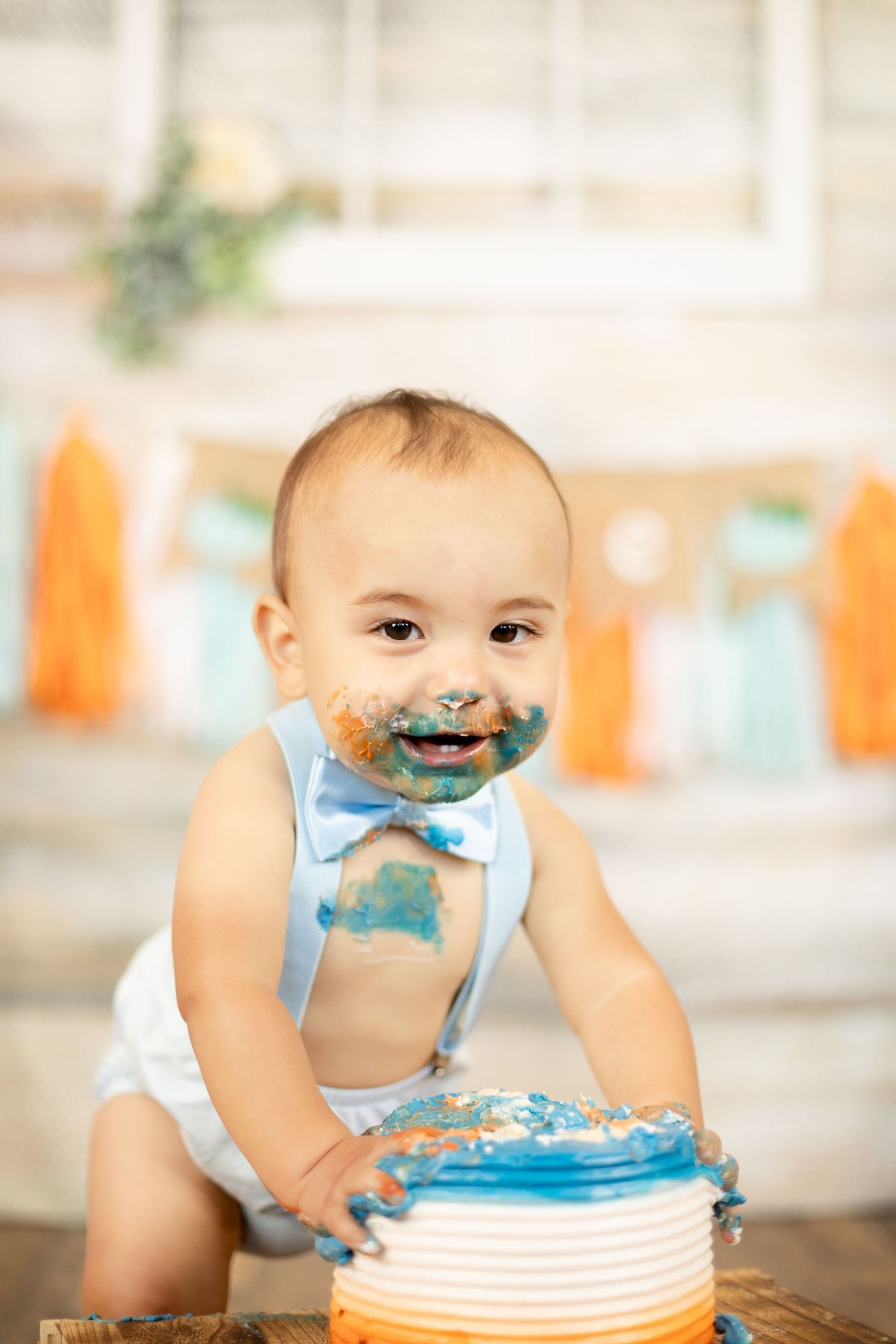 Baby boy with blue frosting on his face smashing his first birthday cake wearing a blue bow tie outfit.