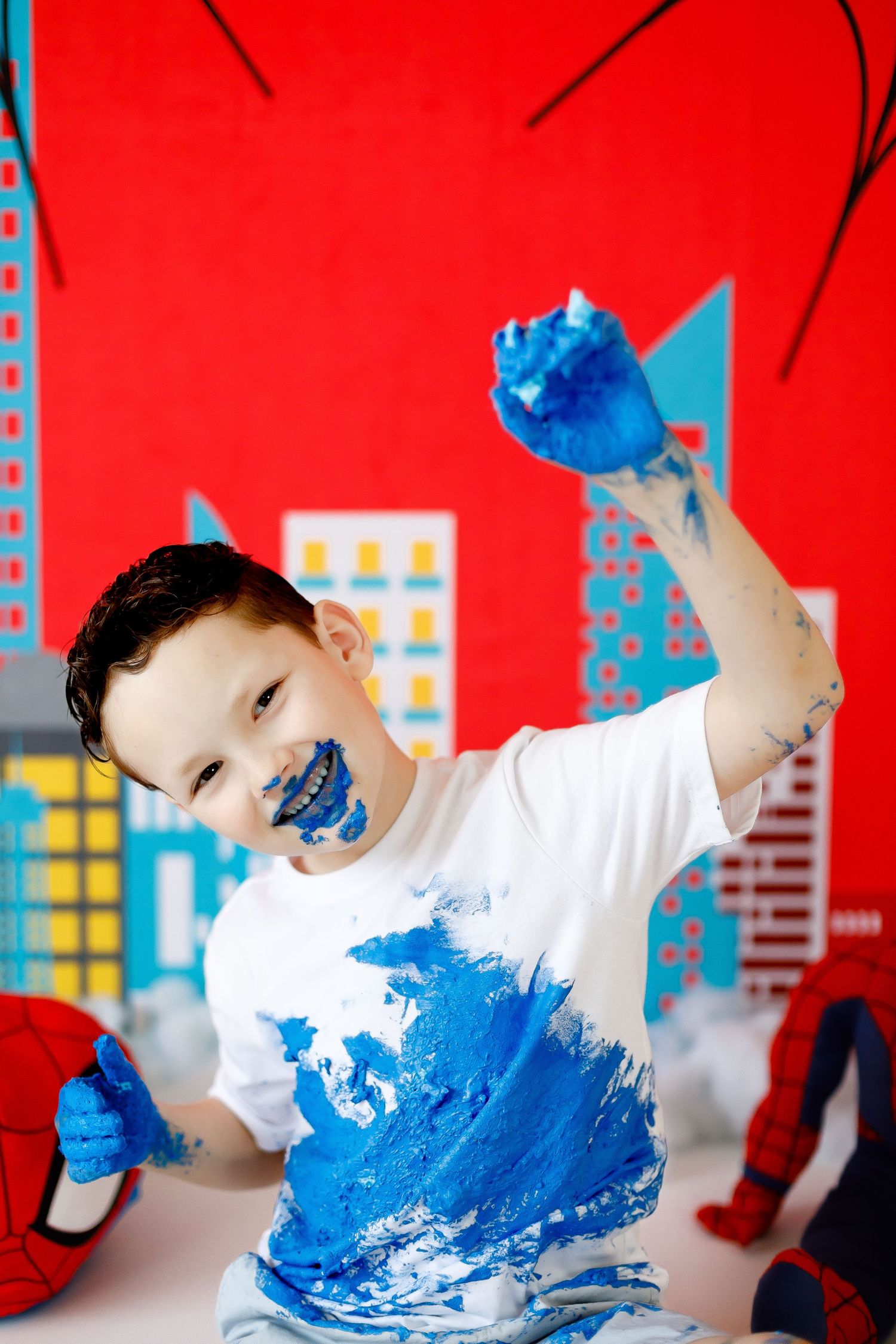 An excited boy raises his blue frosting-covered hands at a Spiderman-themed cake smash party.