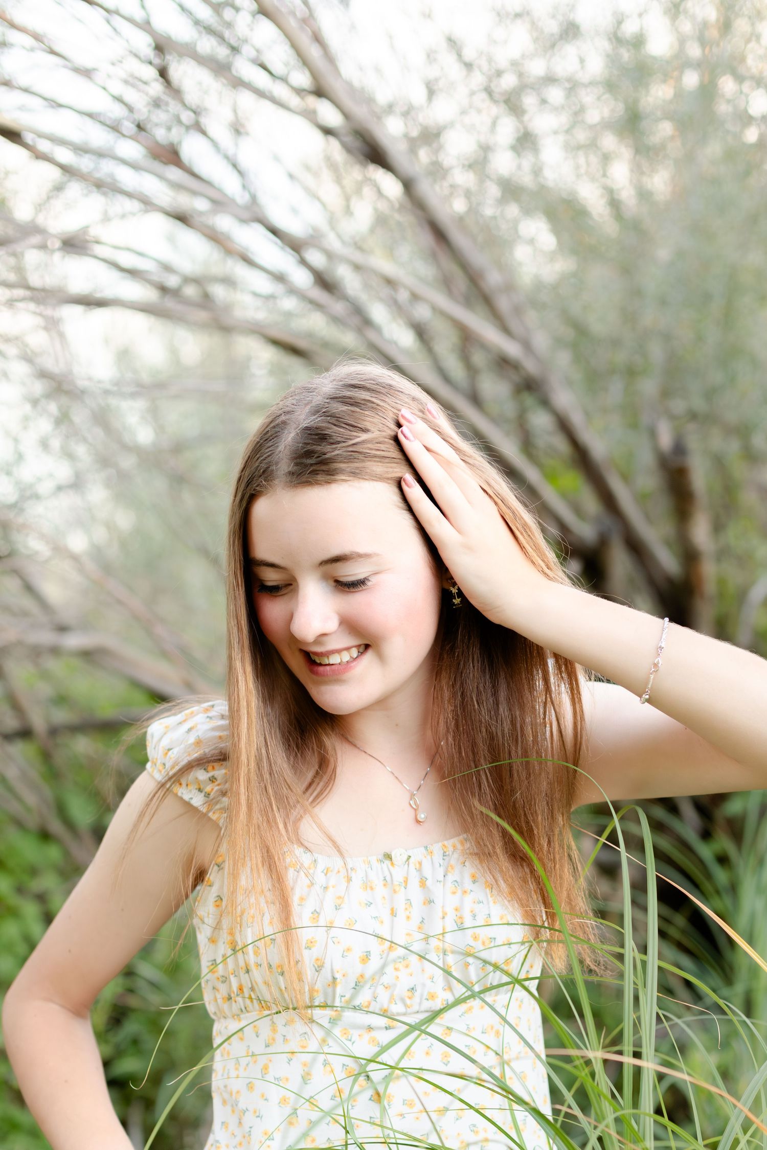 Smiling teenage girl in floral sundress touching her hair while standing among tall grasses and trees.