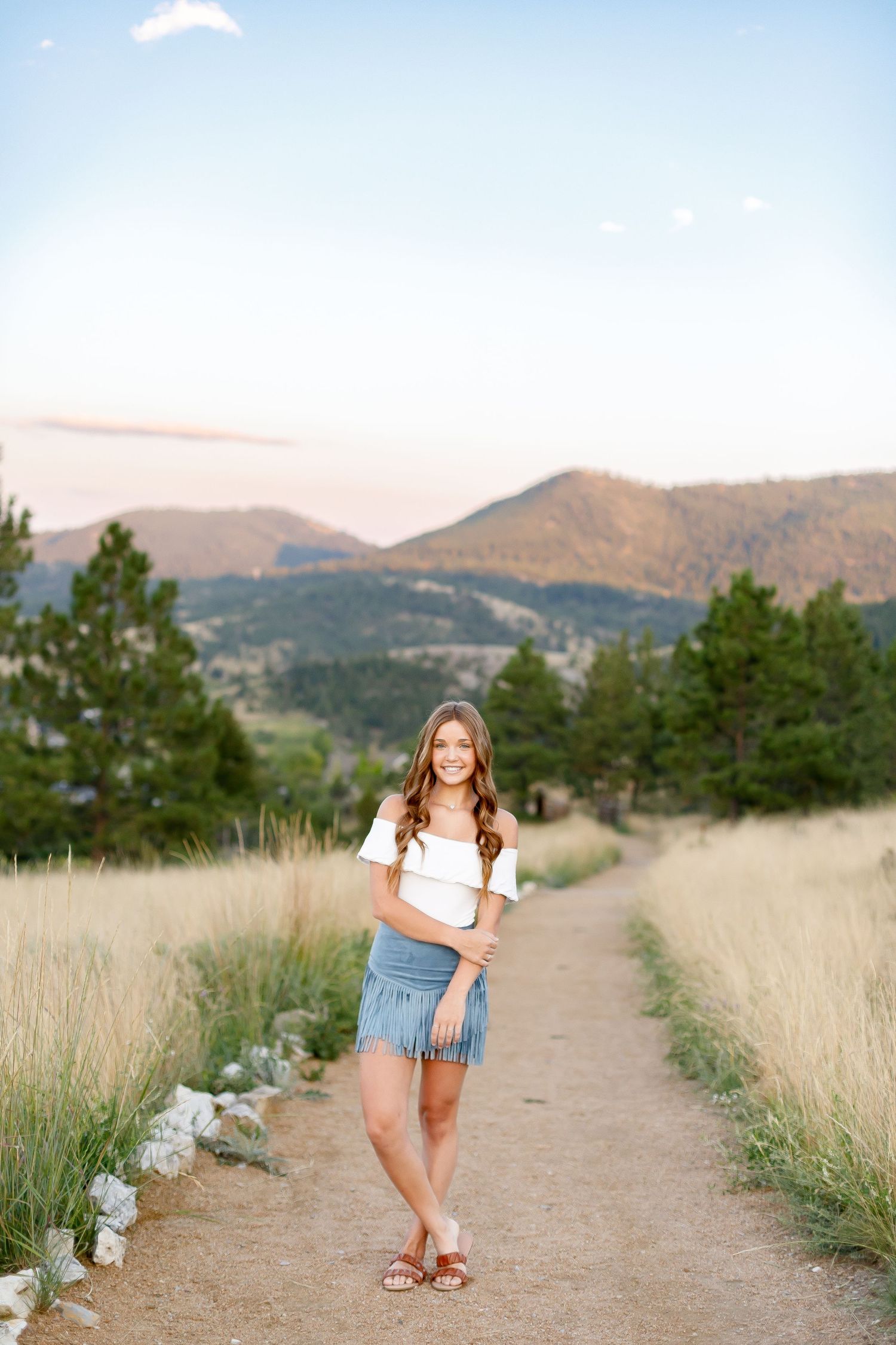 Young woman in white off-shoulder top and denim skirt posing on mountain trail at sunset.