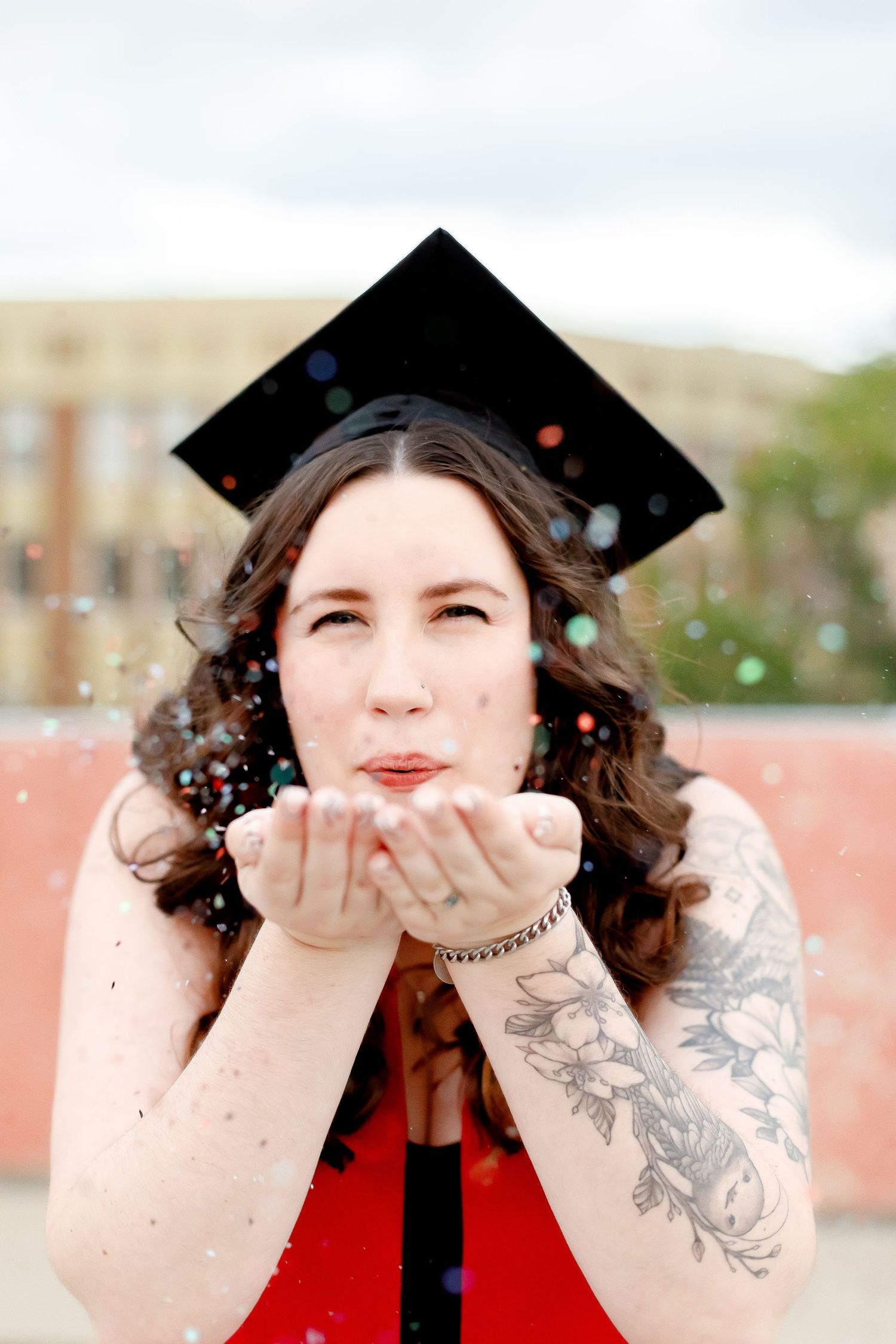 Graduate in red dress and mortarboard blowing colorful confetti from her hands toward the camera.