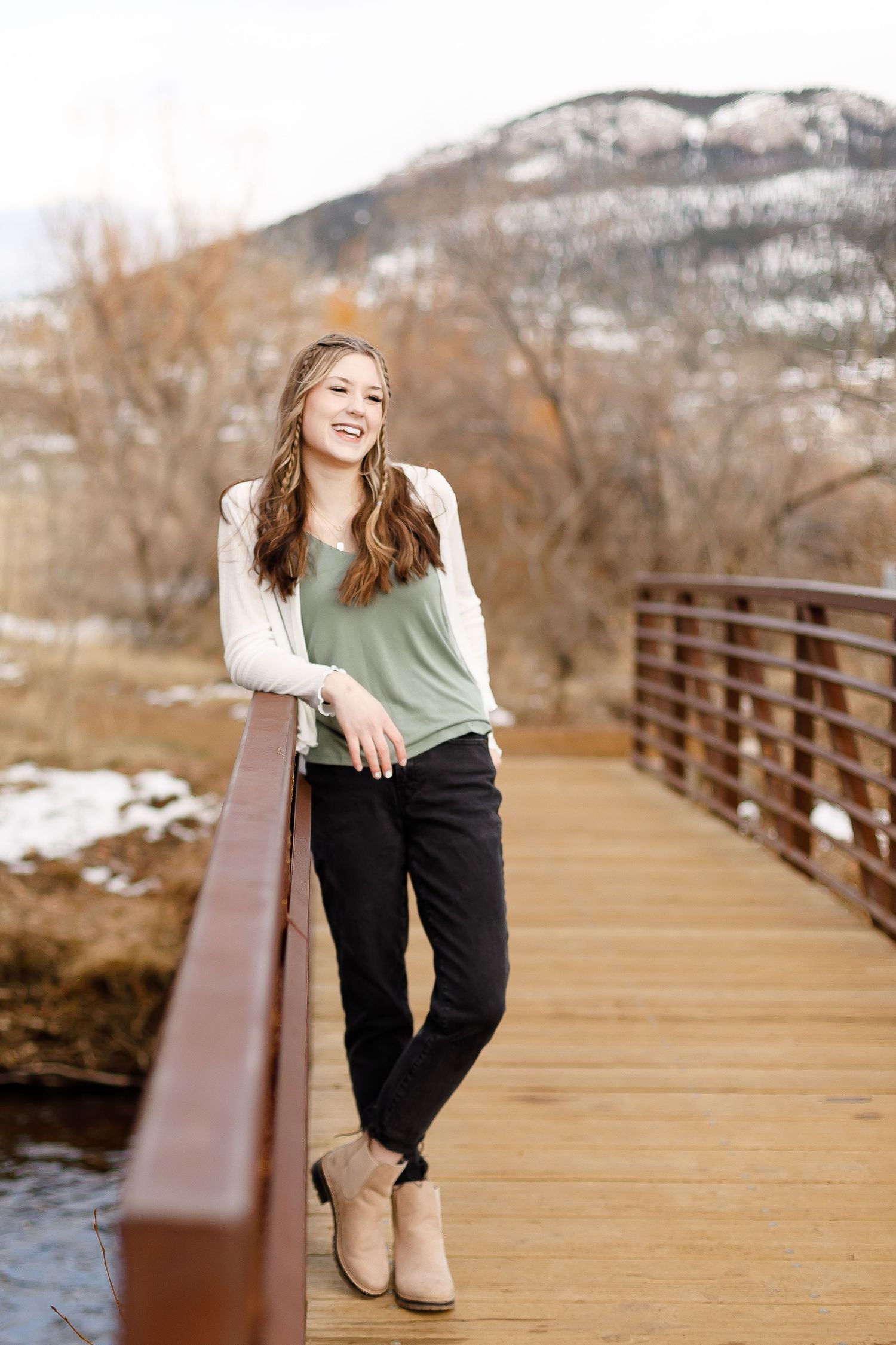 Smiling teenage girl in sage green top leaning on a wooden bridge railing with snowy mountains.