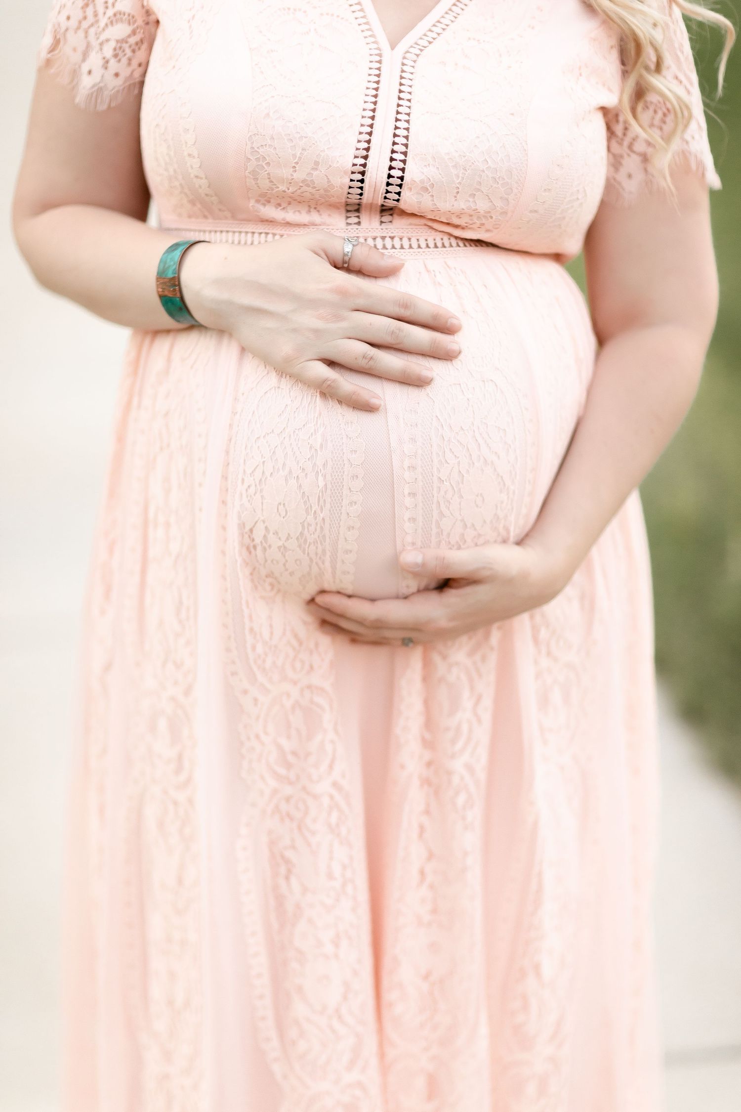 Close-up of pregnant woman in pink lace dress cradling baby bump with both hands.
