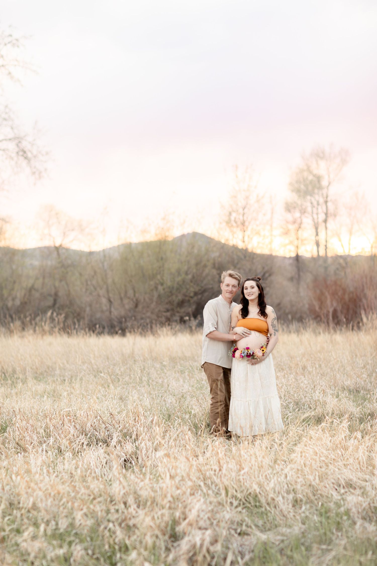 Expecting couple embraces in golden field at sunset, woman wearing orange crop top and white skirt.