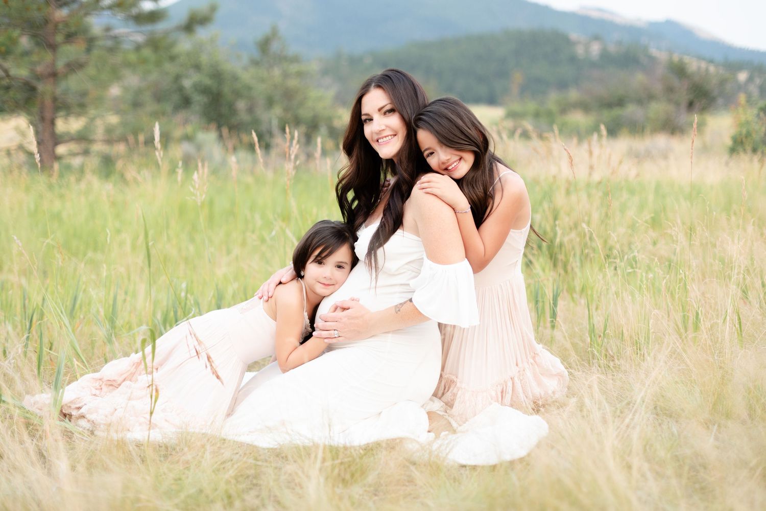 Maternity photo of brunette mom sitting in tall grass with two young daughters.