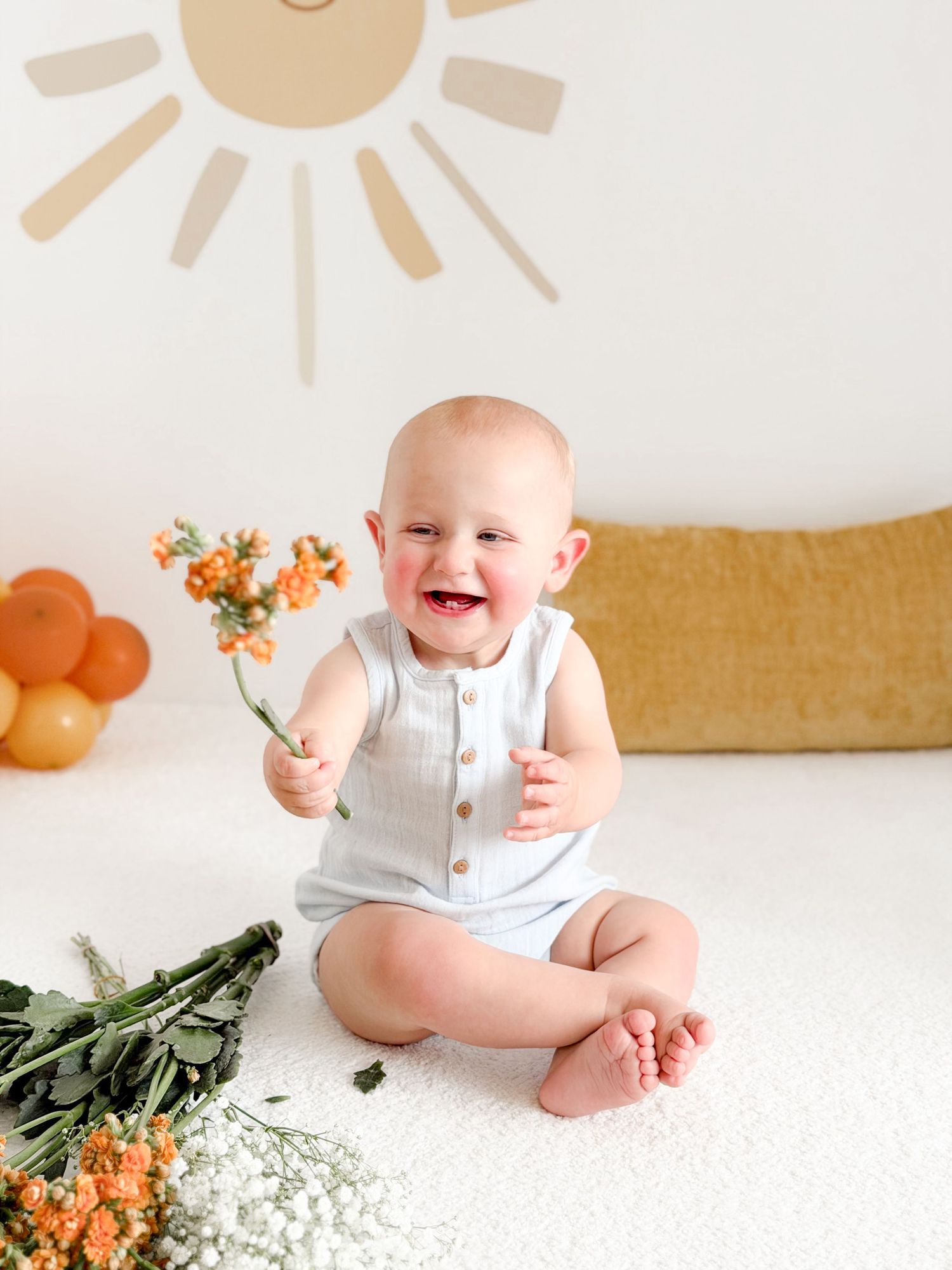 Joyful baby boy sitting on white rug holding orange flowers with sun mural behind him.