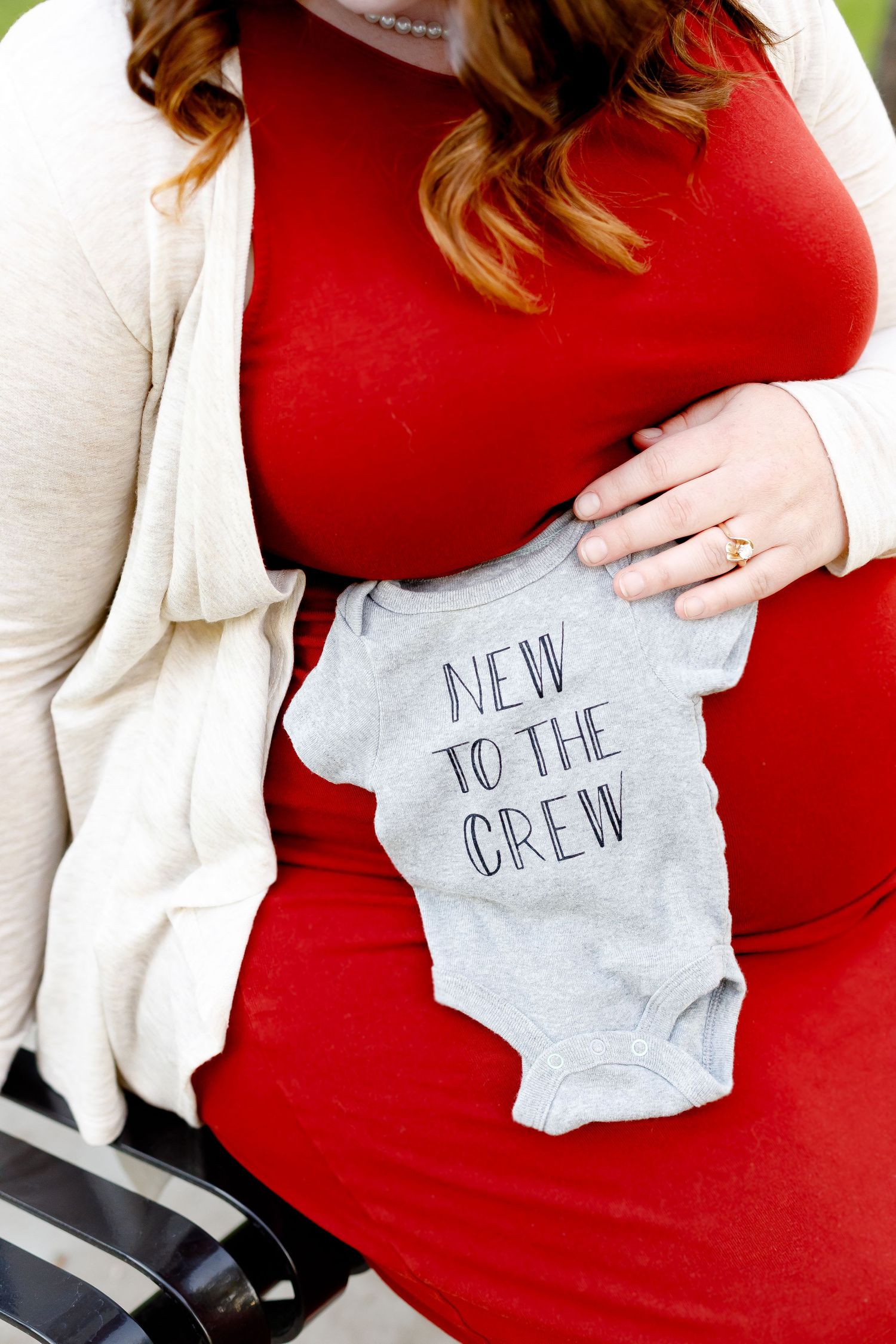 Pregnant woman in red dress holding gray New to the Crew baby onesie against her baby bump.