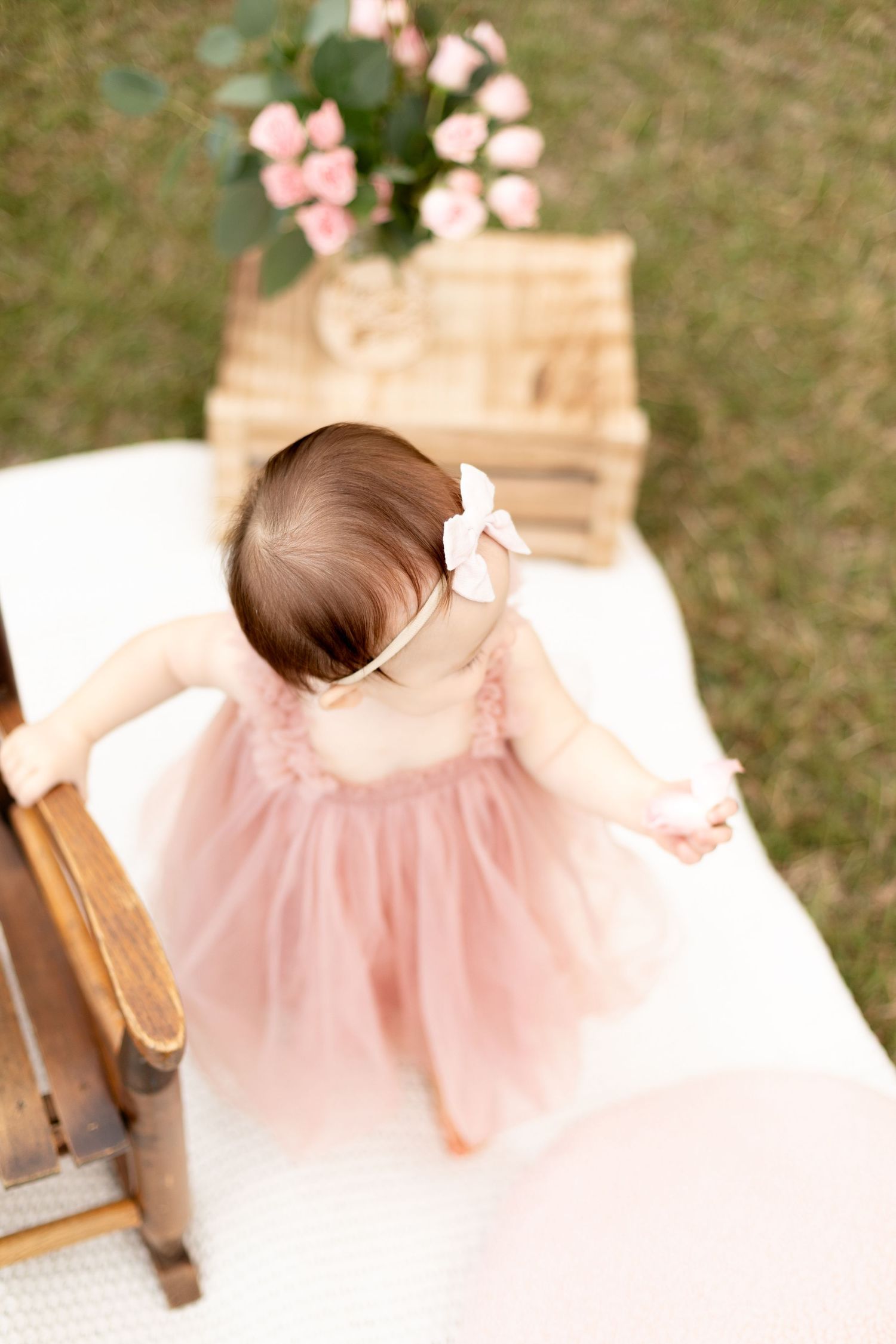 Aerial view of baby girl in pink tulle dress standing at wooden chair with roses in wicker basket outdoors.