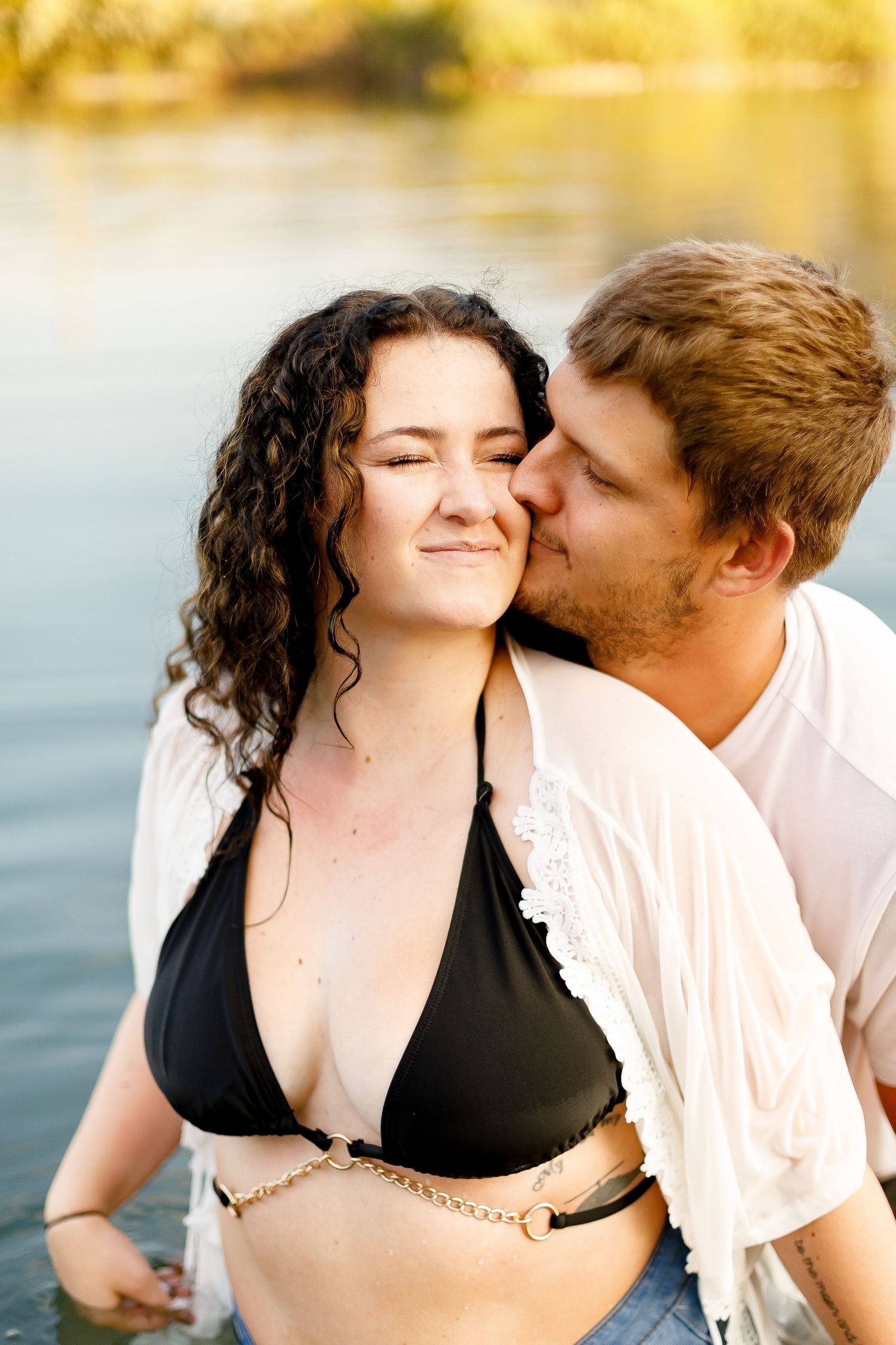 Man kissing woman on cheek while standing in lake, woman wearing black bikini top and white shirt.