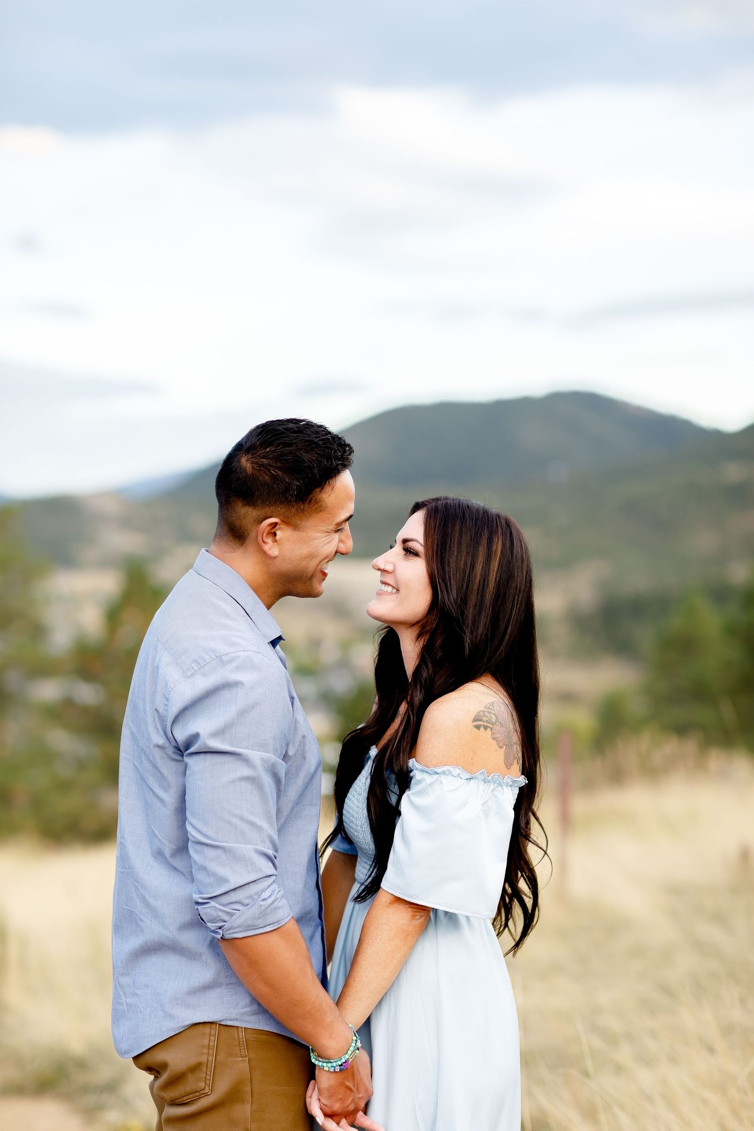 Couple holding hands and smiling at each other in a scenic mountain meadow setting.