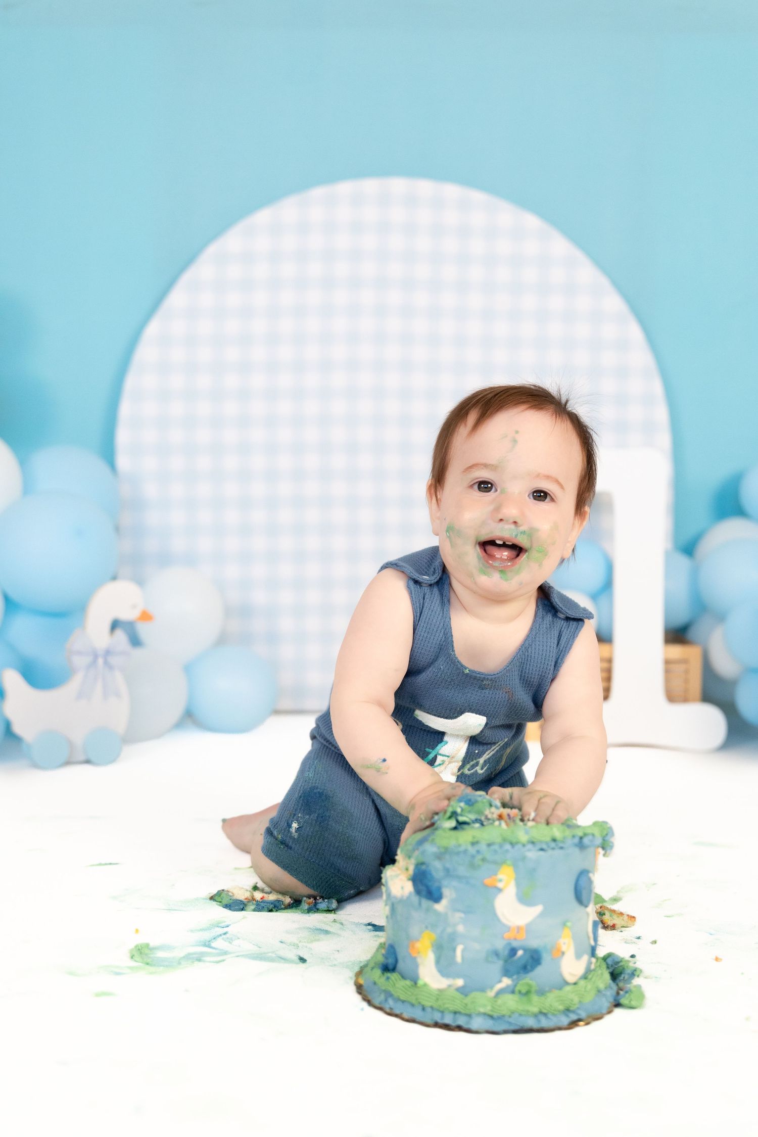Joyful baby boy with cake-smeared face crawls near partially destroyed blue duck first birthday cake.