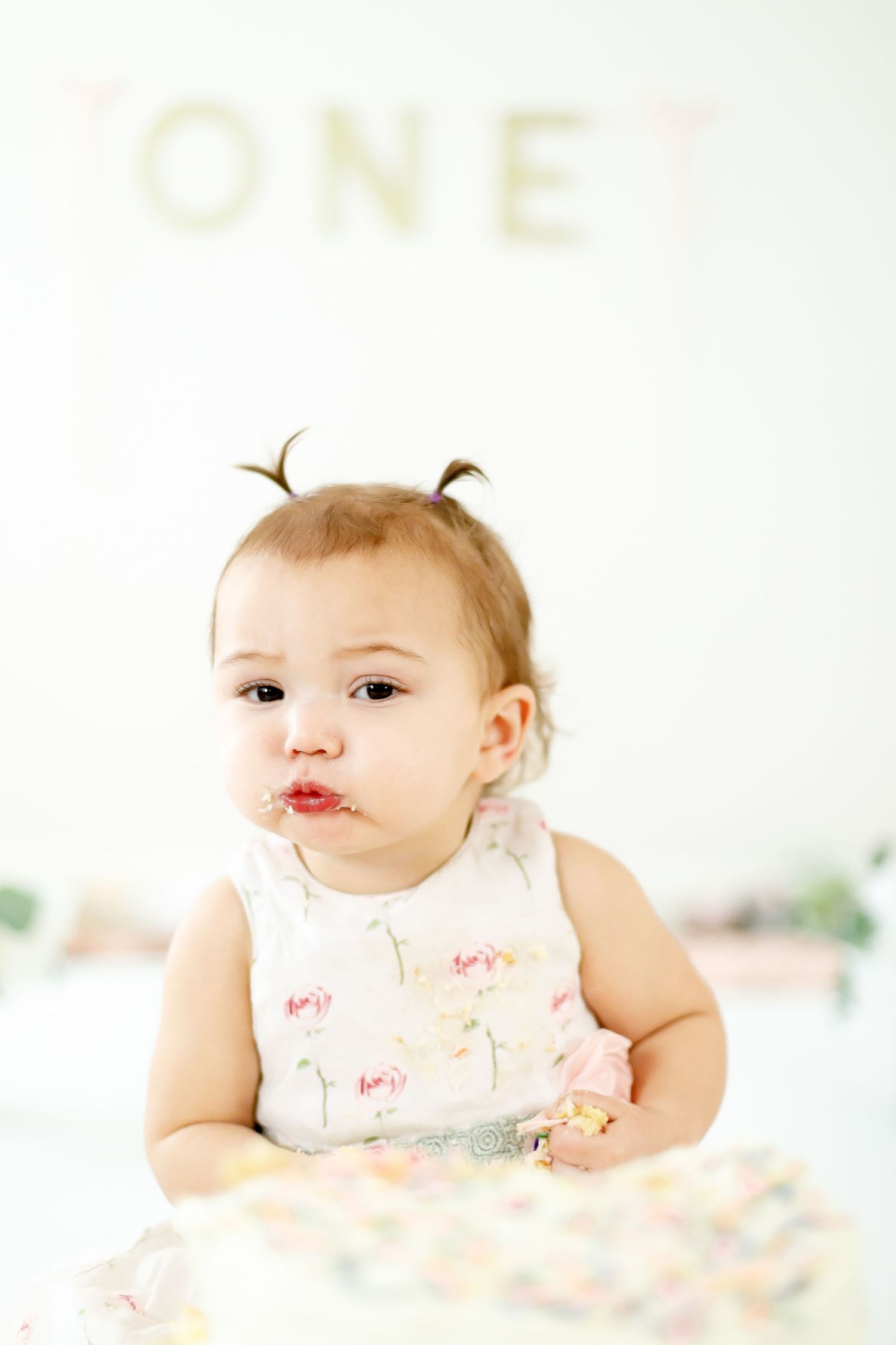 A baby girl with tiny pigtails enjoys her first birthday cake smash, with frosting on her lips.