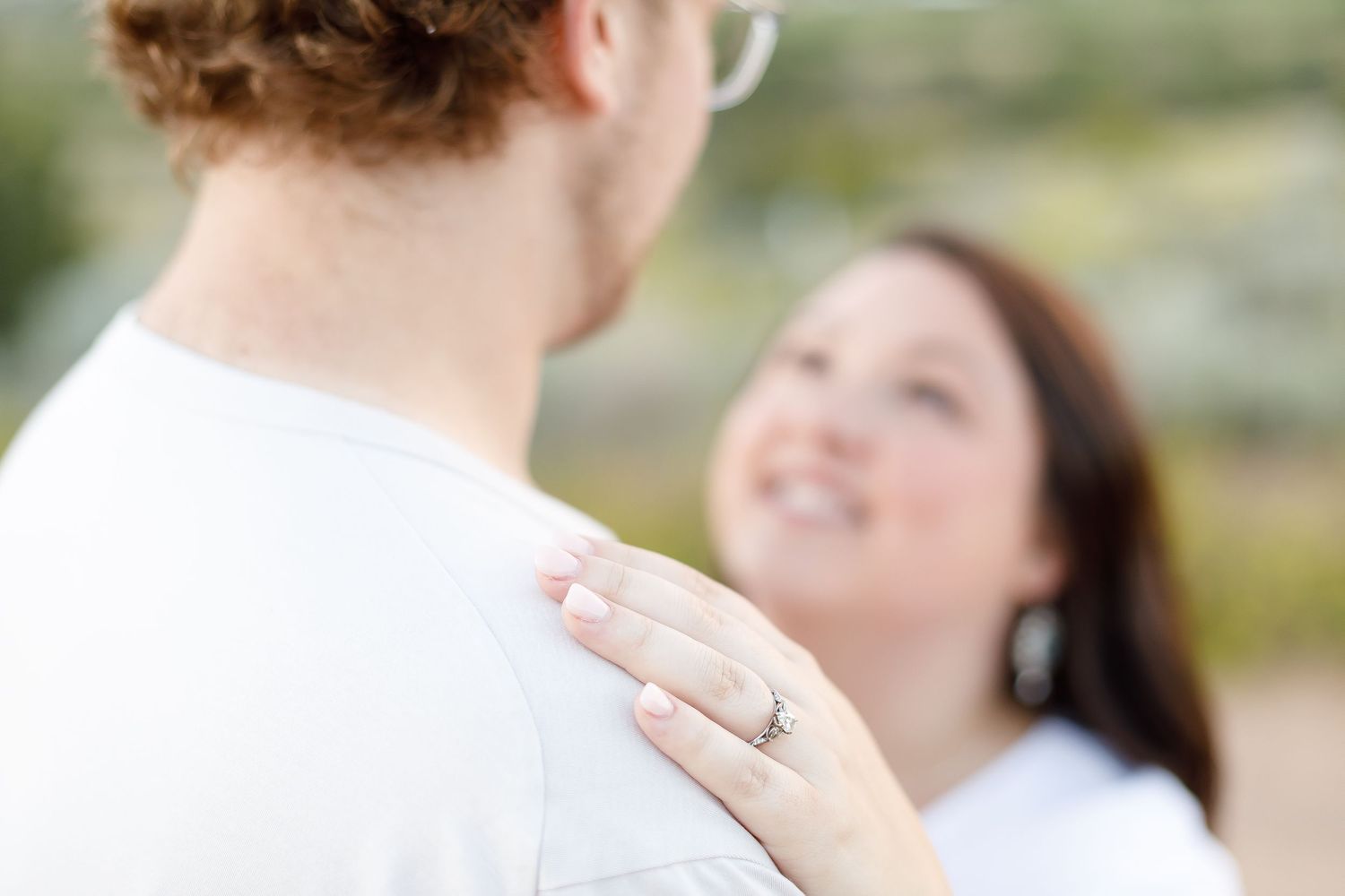 Close-up of a woman smiling at her partner, showcasing her engagement ring on his shoulder outdoors.
