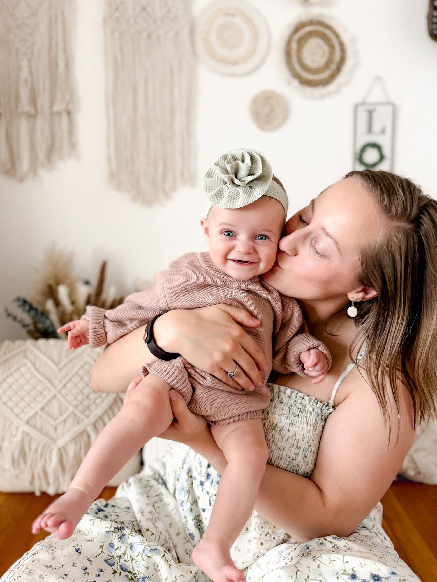 Mother kissing smiling baby wearing green bow headband in boho decorated room.