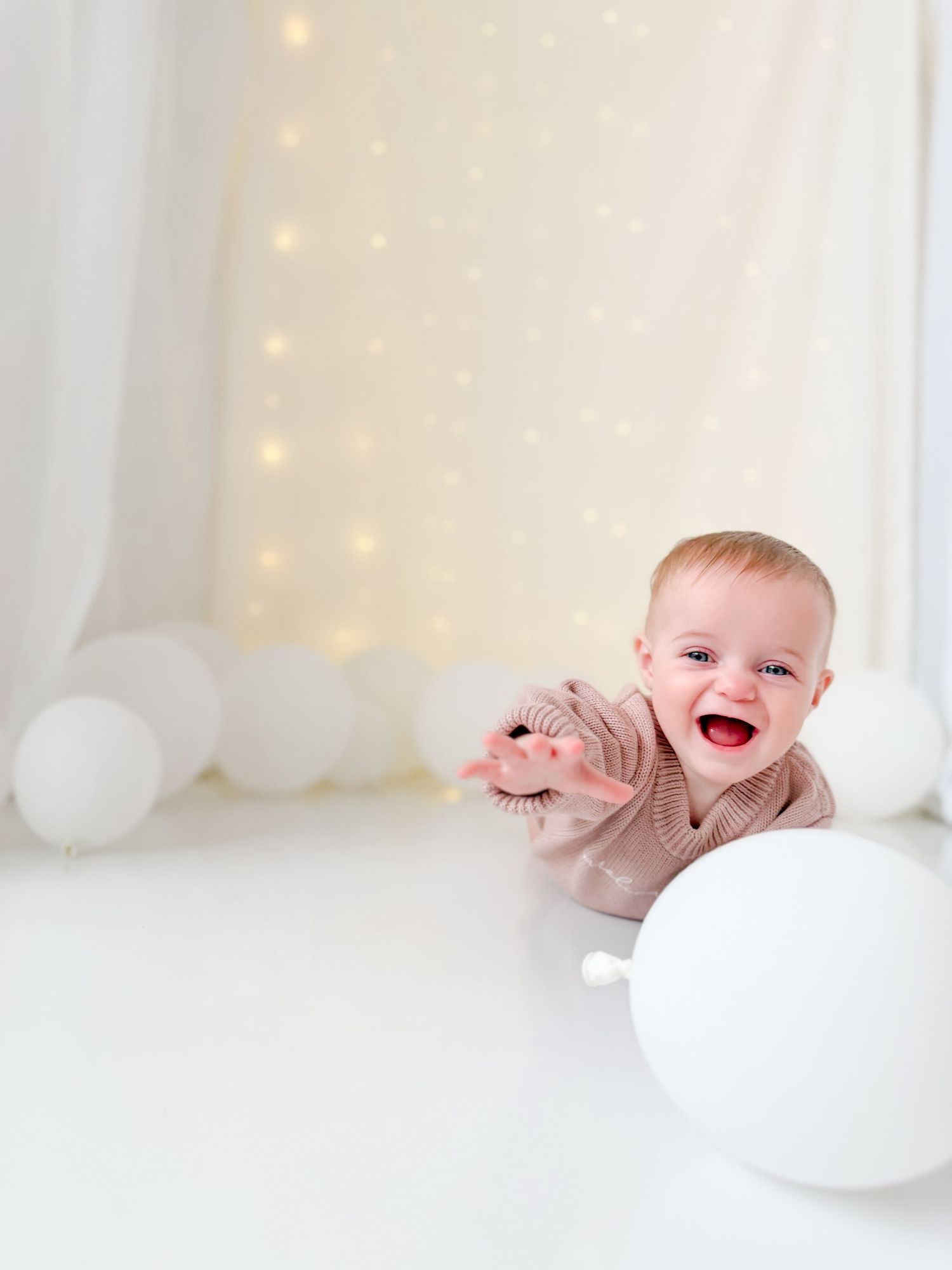 Happy laughing baby crawling toward white balloons in front of twinkling light backdrop.