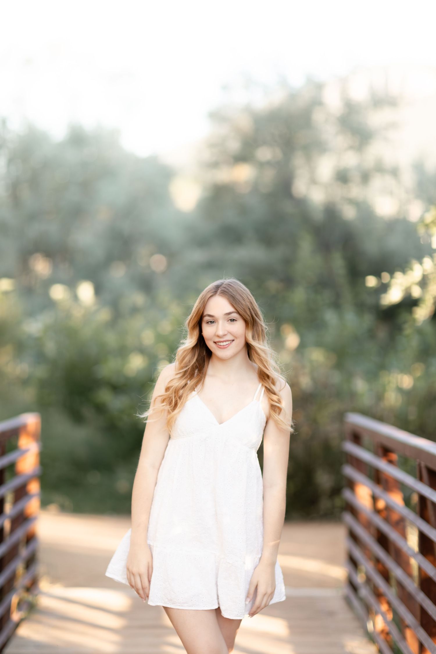 Young woman in white romper smiling on wooden bridge surrounded by lush green trees in golden hour light.