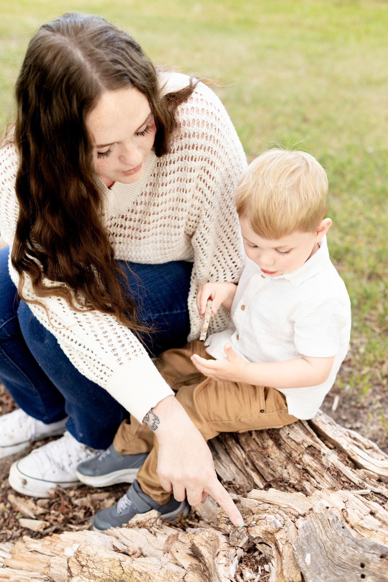 Mother and young toddler son exploring a fallen log together in a park setting.