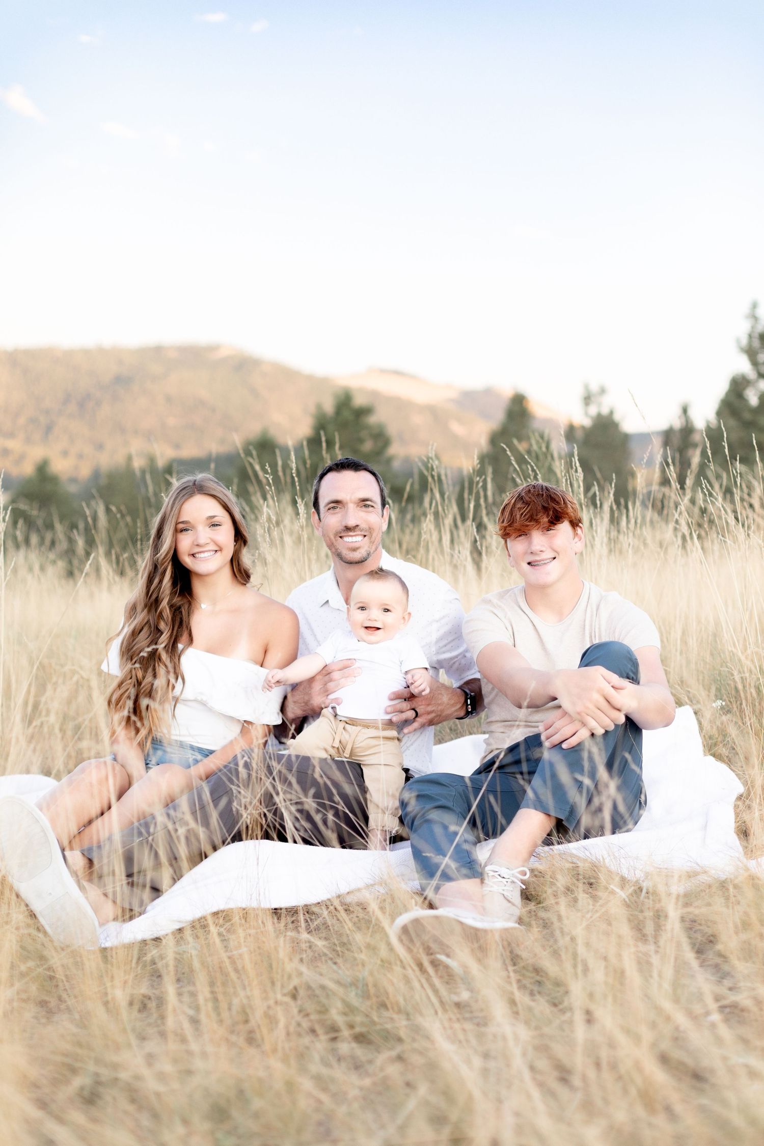 A family of four sits on a white blanket in a golden grass field with mountain views behind.