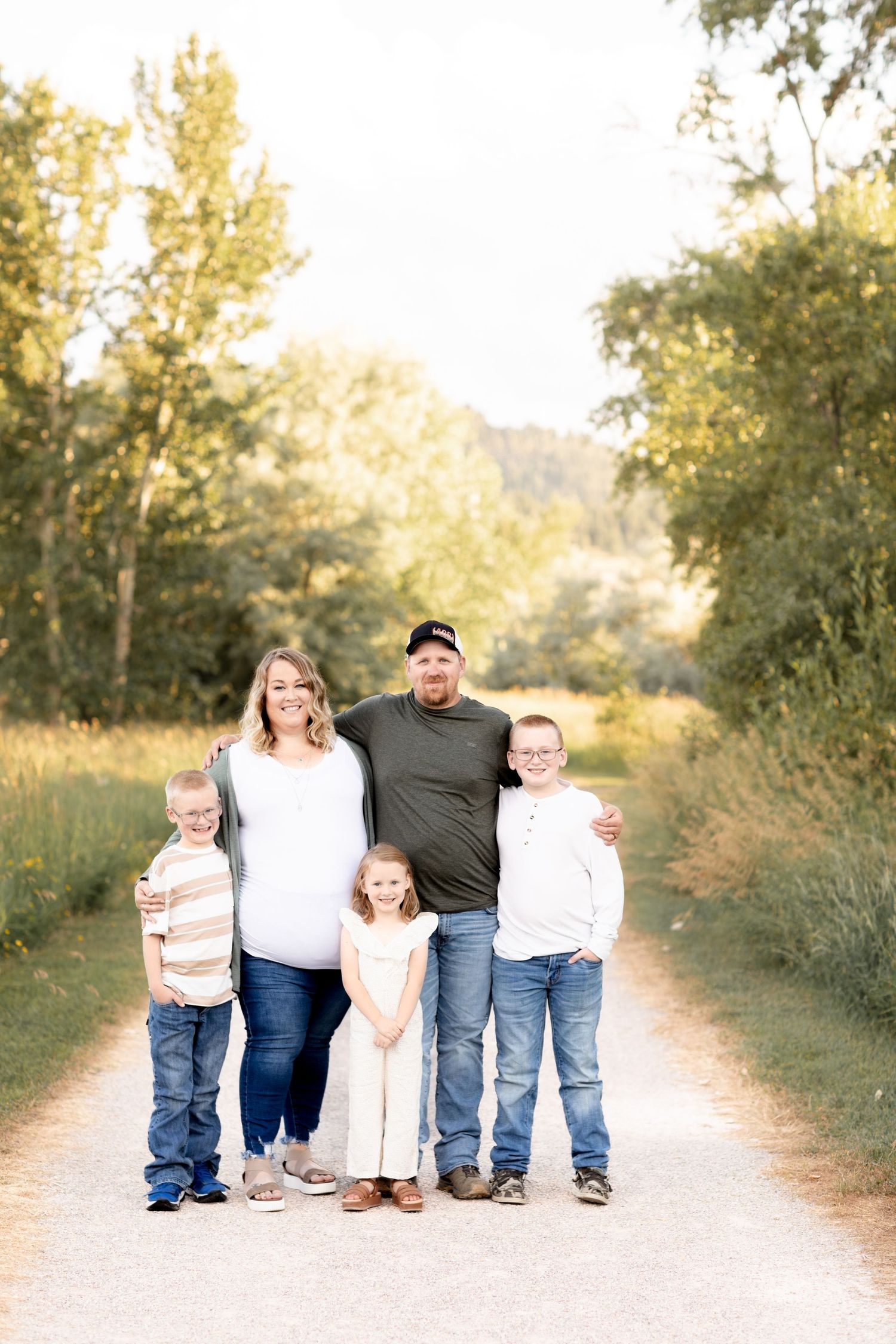 Family of five with pregnant mother posing on a tree-lined path in golden hour light.