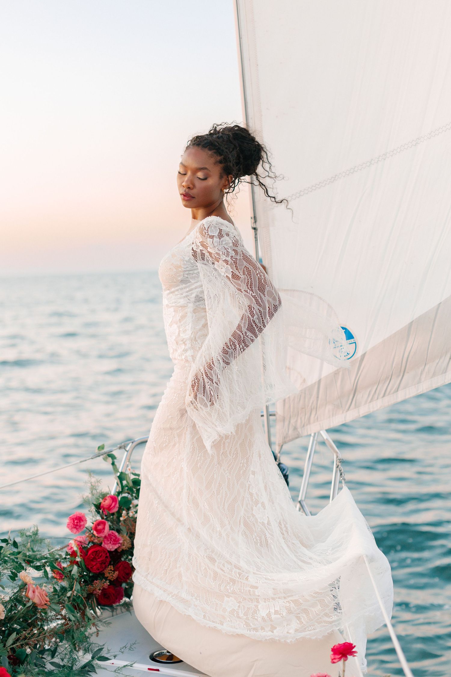 Elegant Black bride in flowing lace gown standing on sailboat at sunset over ocean.