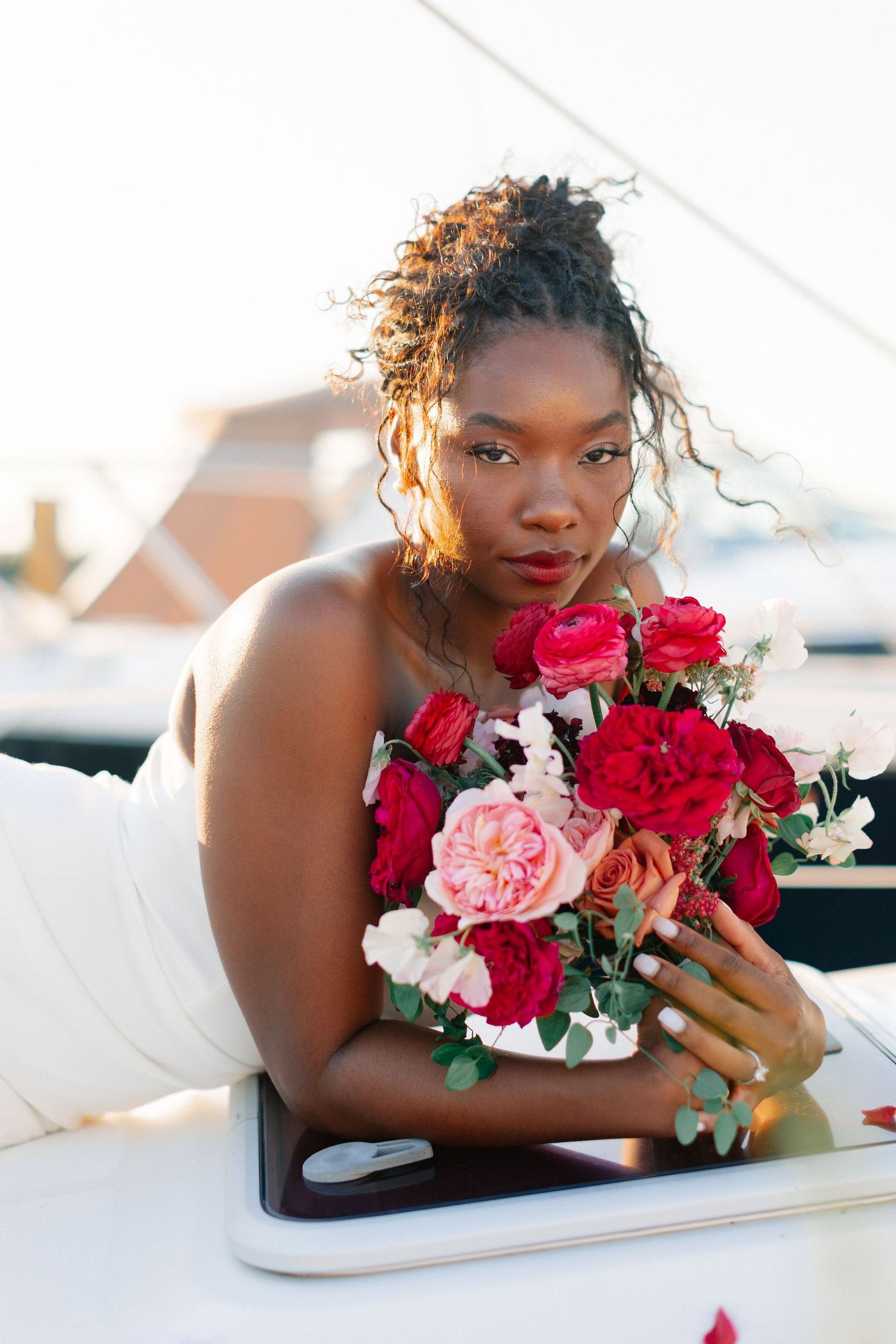 Elegant Black bride in white dress holding lush red and pink rose bouquet on a sunlit sailboat.