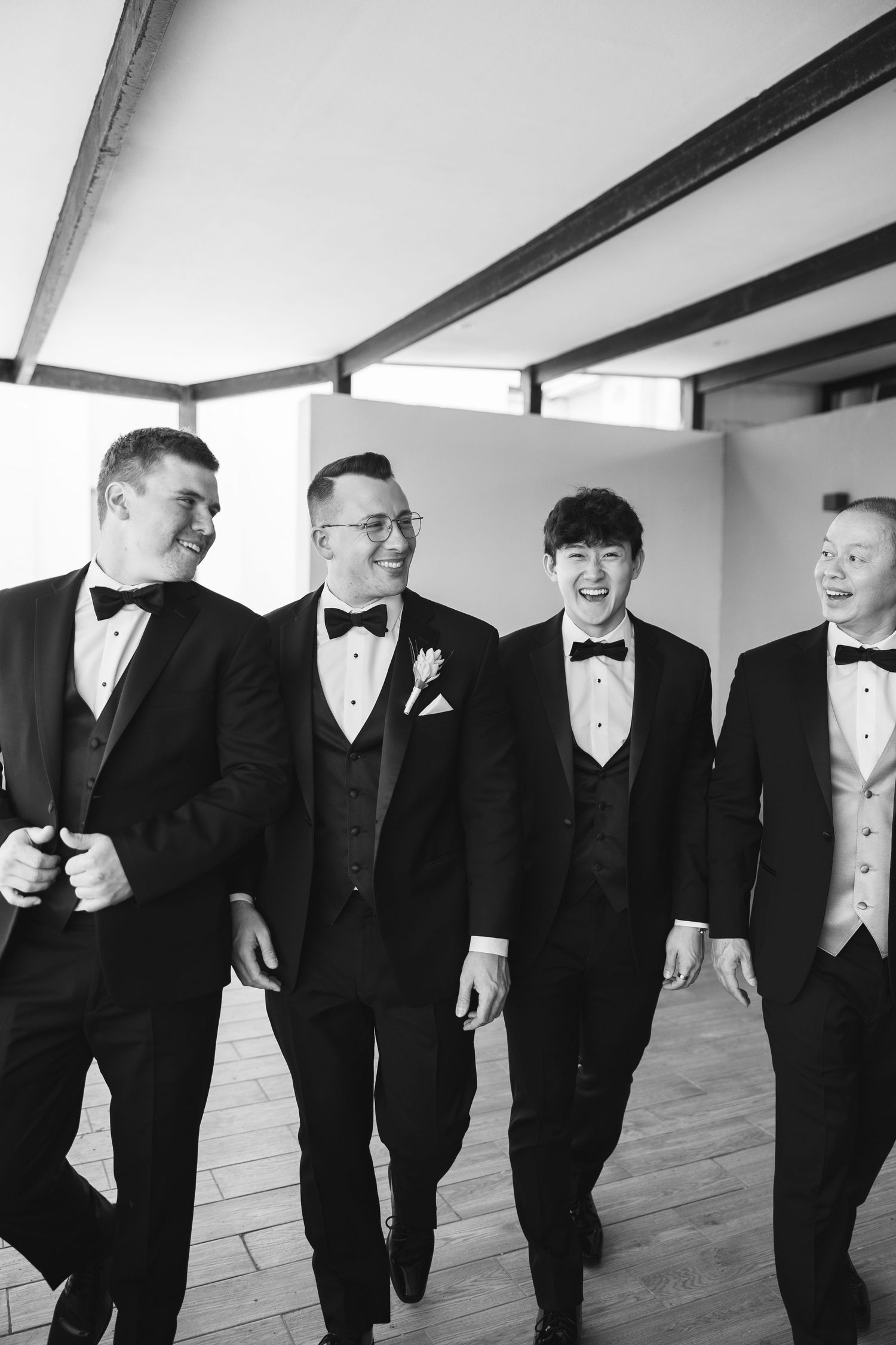 Four groomsmen in black tuxedos and bow ties laughing and walking together in a black and white wedding photo.