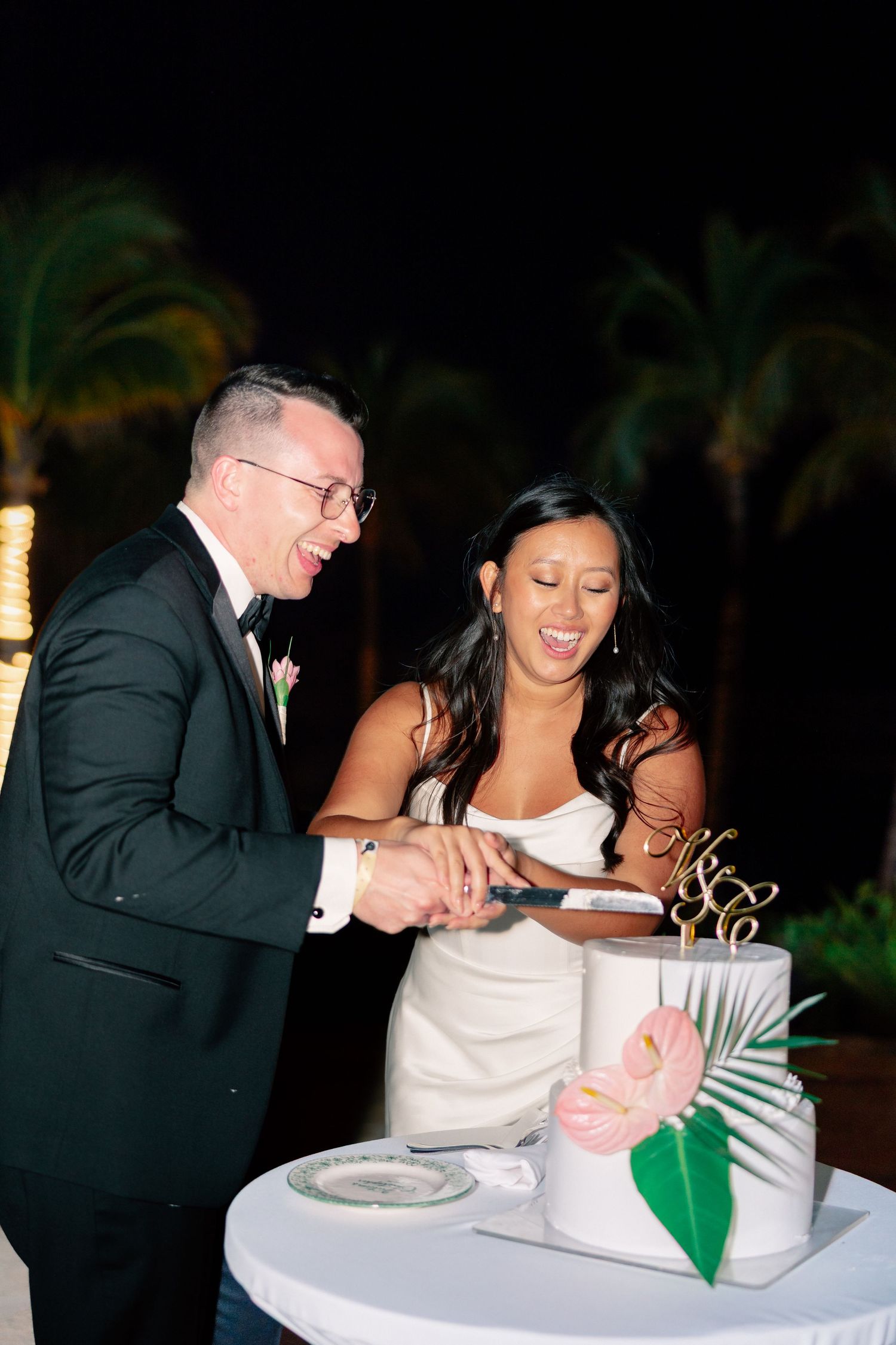 Bride and groom laughing while cutting their tropical wedding cake with palm leaf and pink floral decor at night.