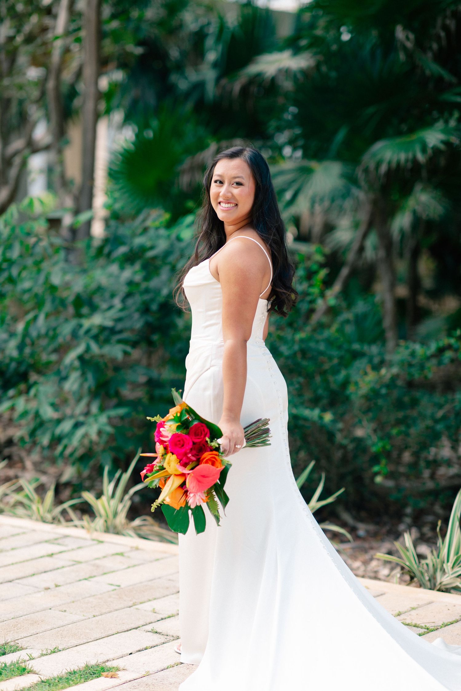 Smiling bride in white gown holds vibrant tropical bouquet on wooden path with lush greenery backdrop.