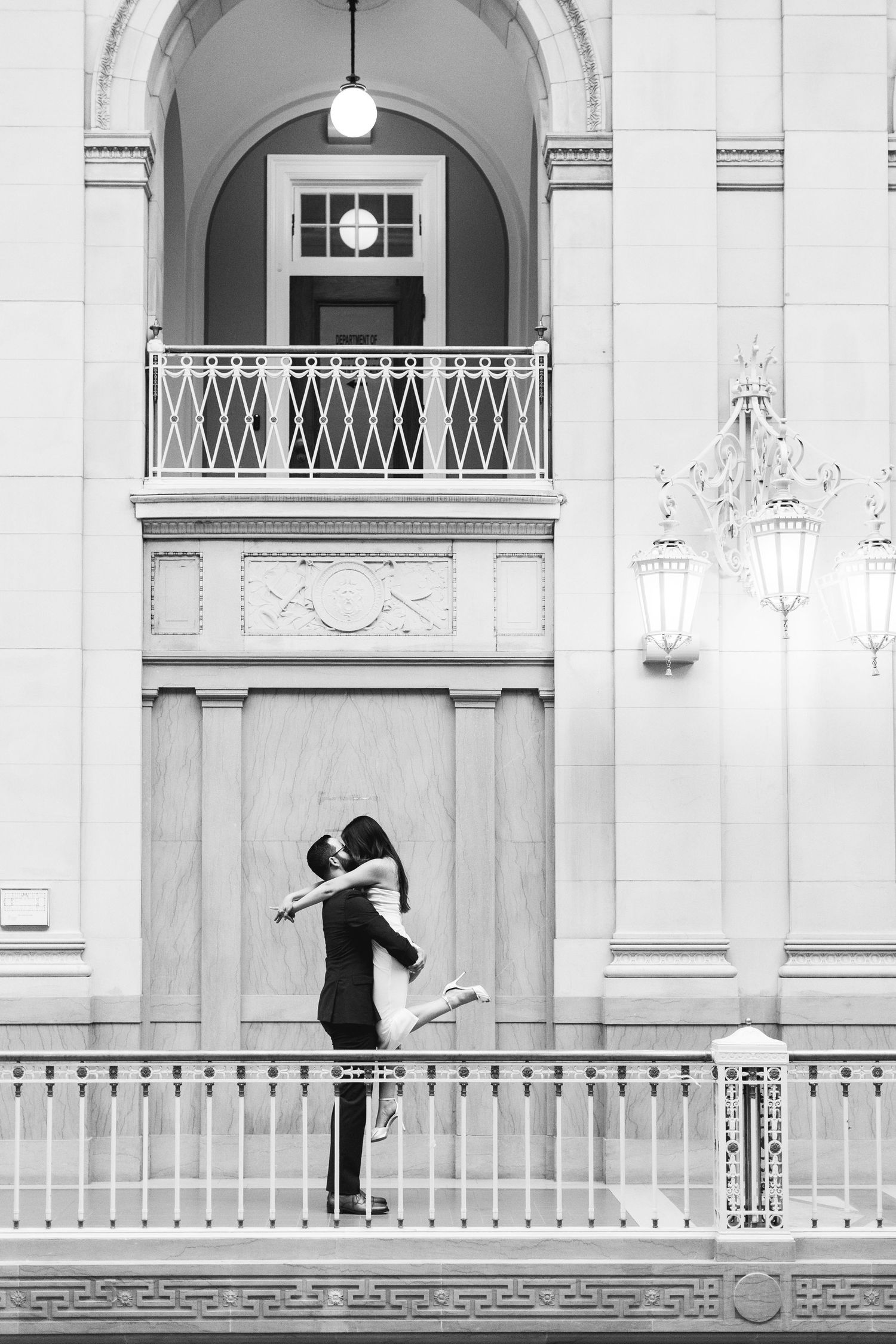 Black and white photo of a couple embracing and kissing in front of an elegant building with arched doorway.