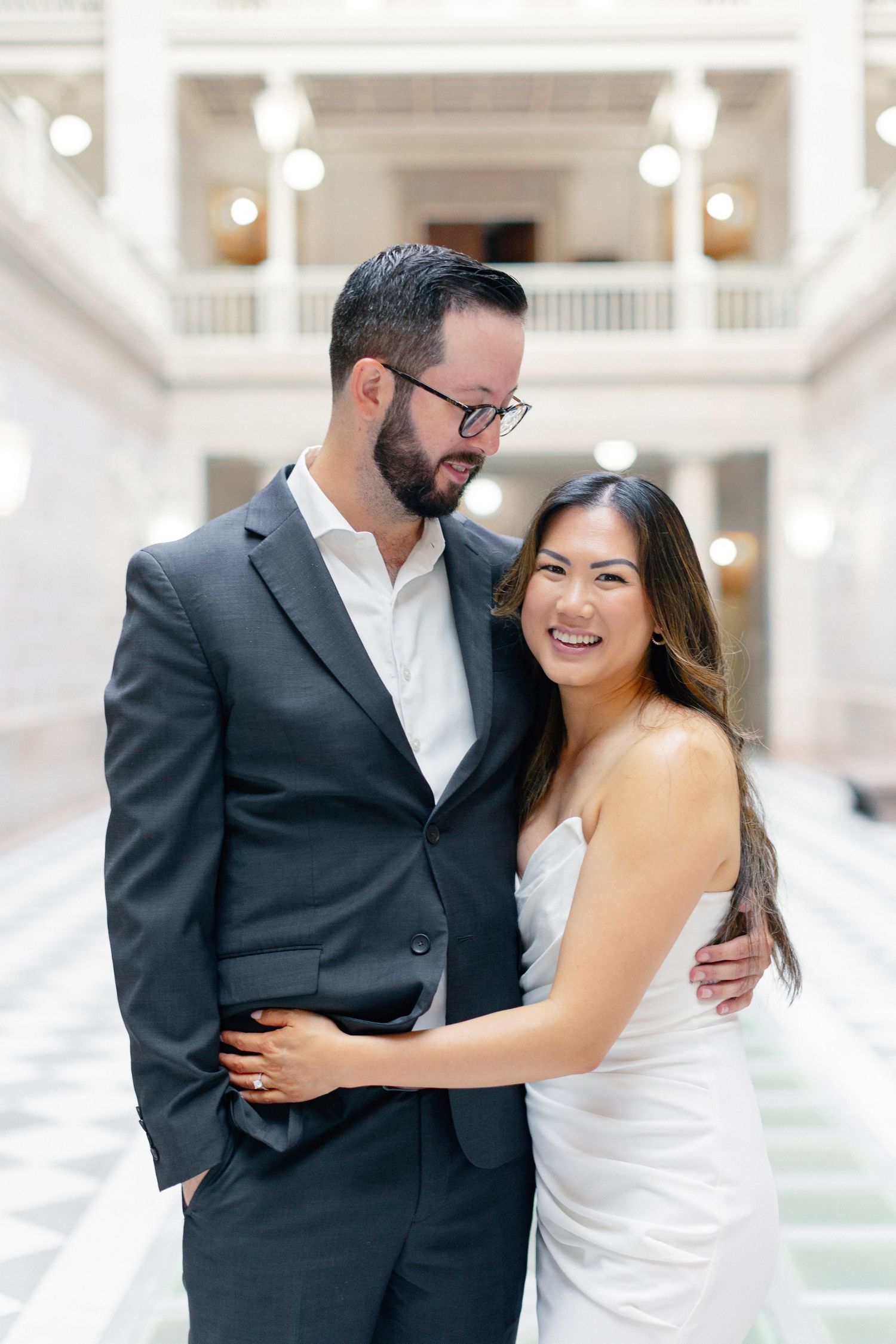 Couple in elegant wedding attire embracing and smiling in a grand white marble hall with ornate balconies.