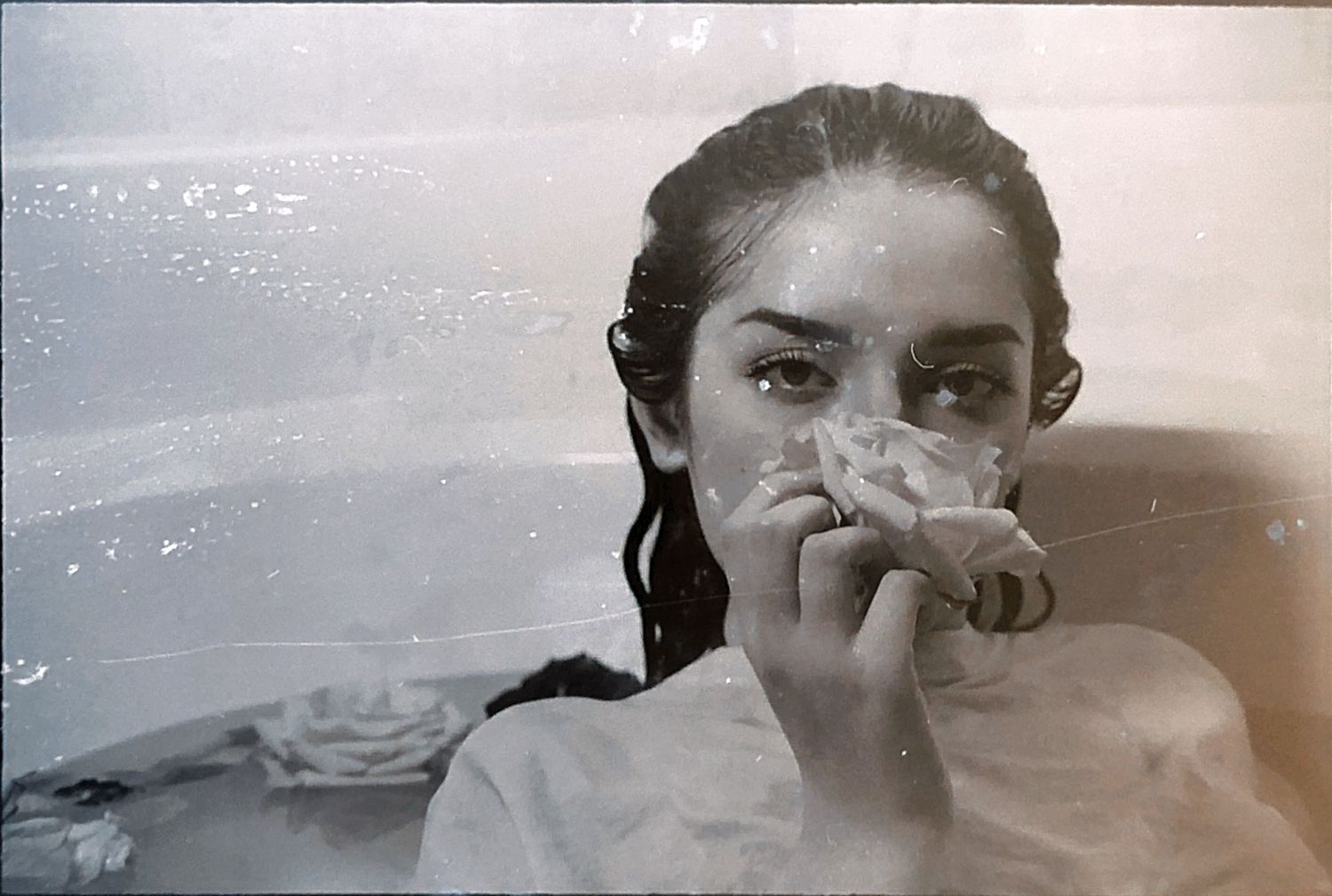 Black and white photo of a young woman in a bathtub, holding a cloth to her face with wet hair.