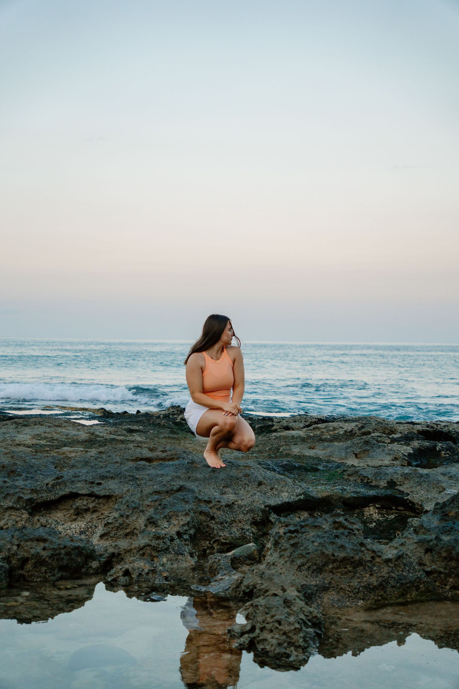Woman in orange top crouching on rocky coastal shore at sunset with ocean waves and reflection in tide pool.