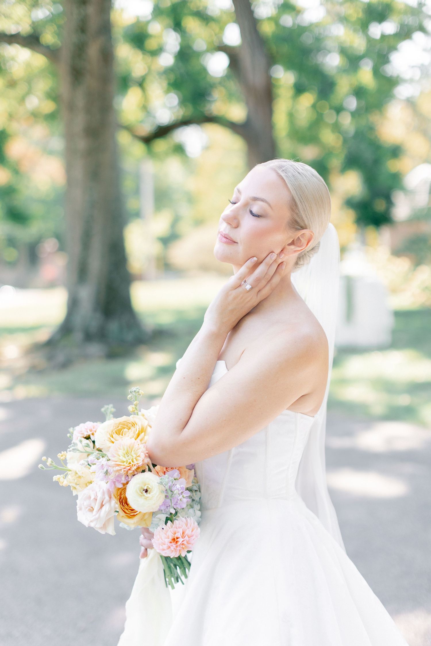 Elegant bride in strapless gown holds pastel floral bouquet with eyes closed outdoors in soft natural light.