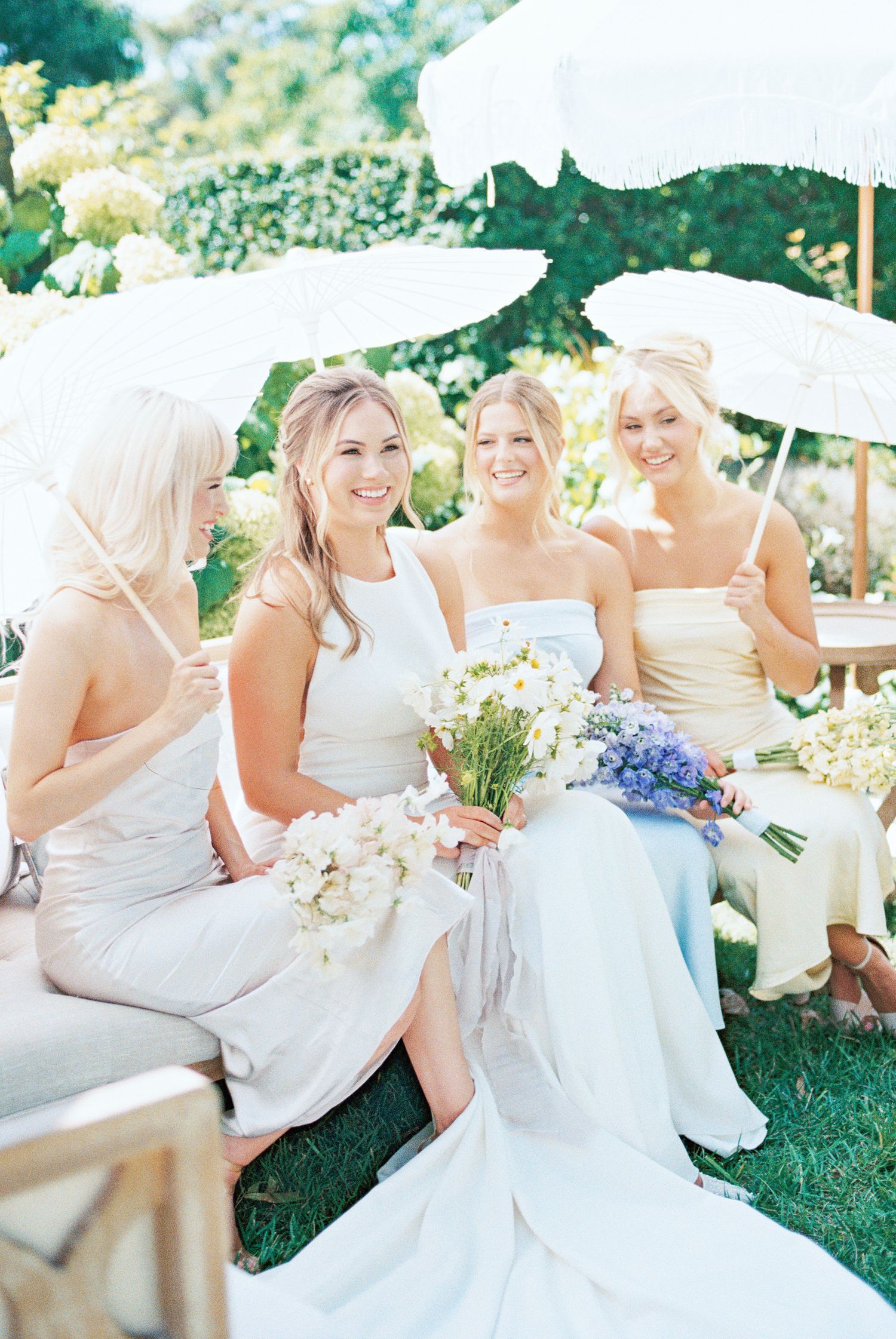 Bride and bridesmaids in white and cream dresses holding floral bouquets under white parasols at an outdoor wedding.
