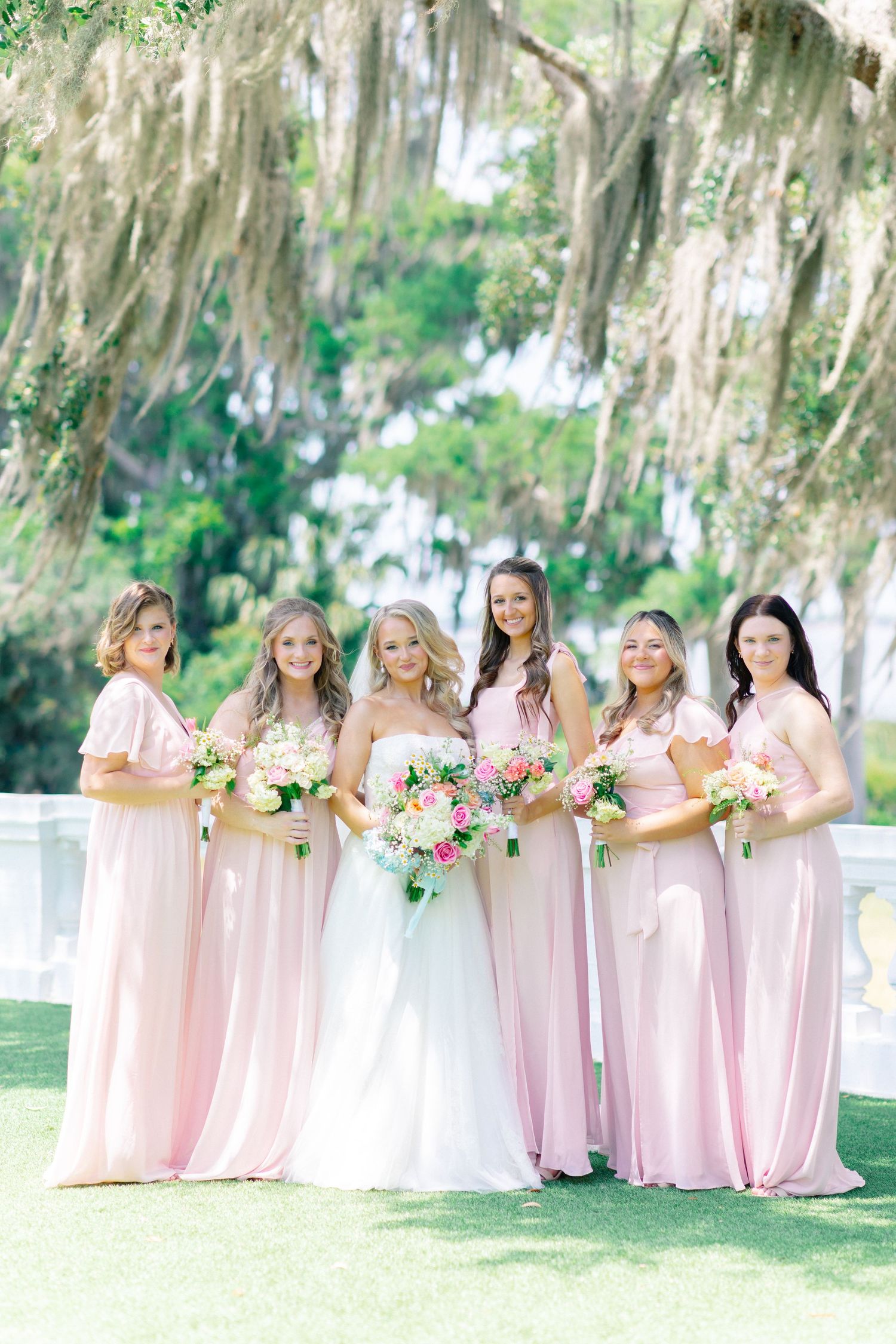 Bride in white gown with five bridesmaids in pink dresses holding floral bouquets outdoors under moss-draped trees, wedding at Bella Cosa
